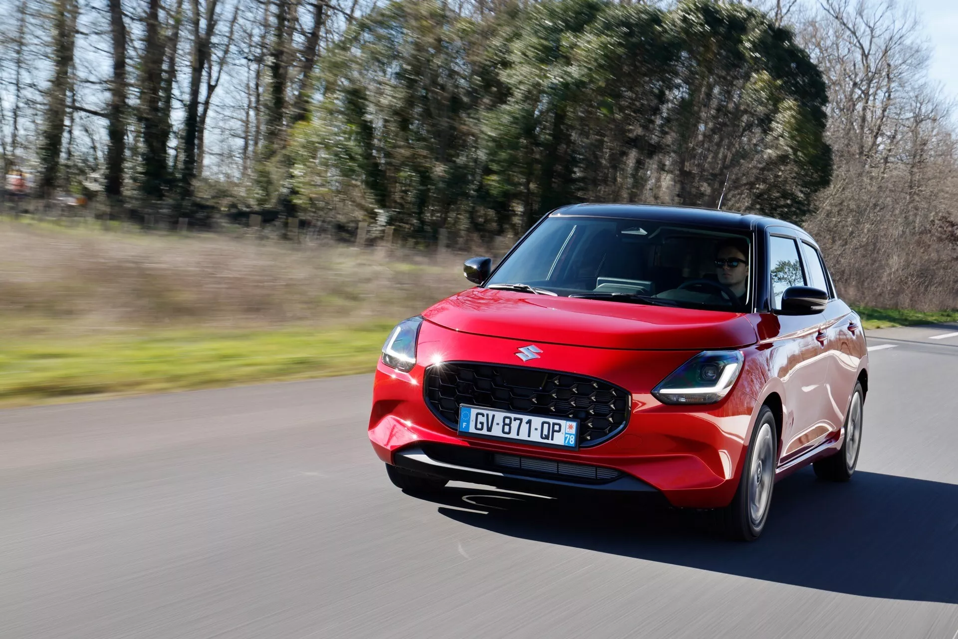 Dynamický záber na červený hatchback Suzuki Swift 2024 v pohybe na asfaltovej ceste so zeleným pozadím, zobrazujúci dizajn prednej ľavej strany vrátane LED svetlometov, mriežky a aerodynamických vlastností za denného svetla