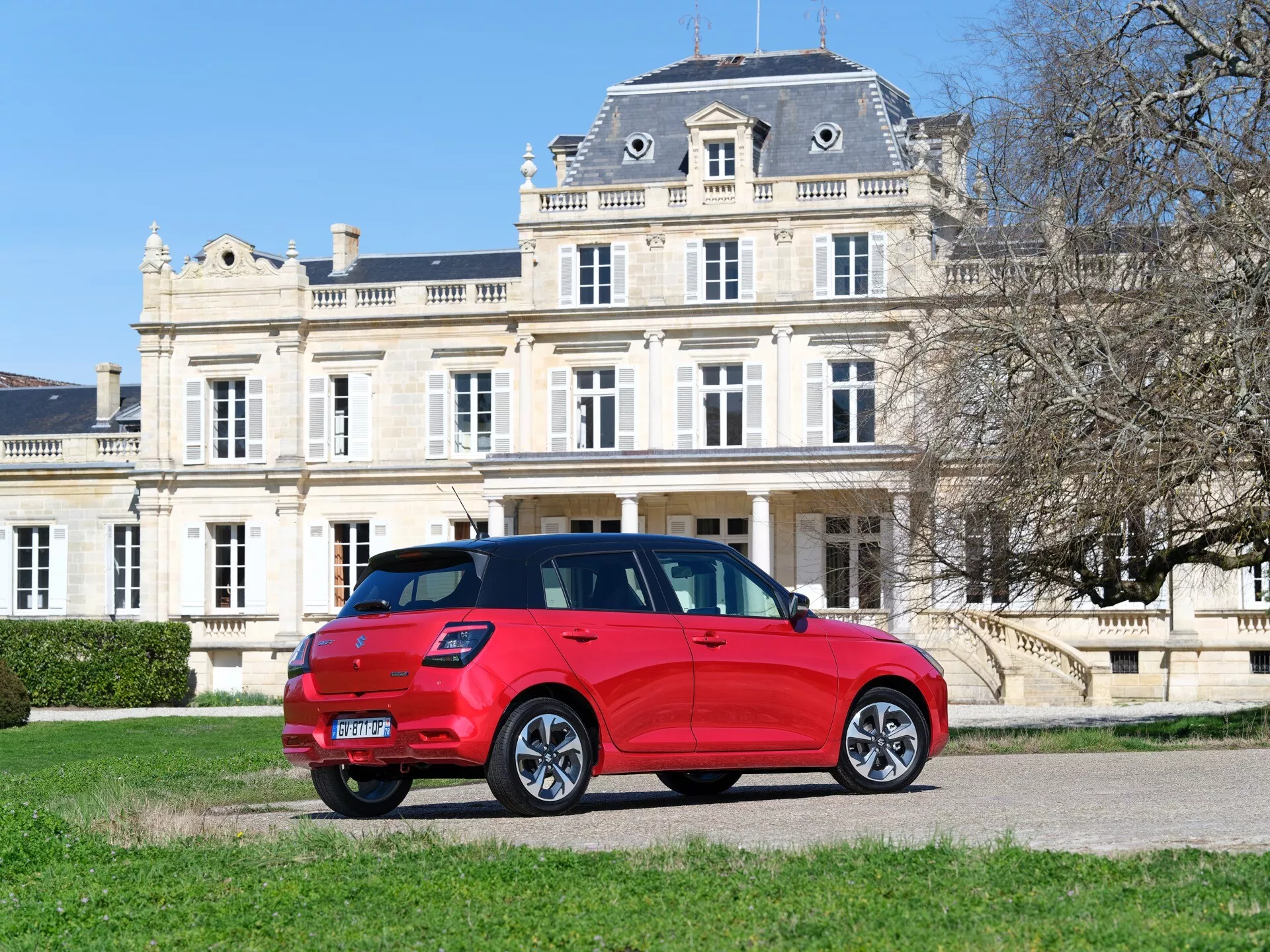 Suzuki Swift 2024, 7. generácia, moderné kompaktné SUV v jasne červenej farbe zaparkované na štrkovej ceste so zeleným trávnikom a zámkom v pozadí, za jasného denného svetla zobrazujúce zadné svetlá, stierač zadného okna, pravé bočné dvere, okná a viaceré zliatinové kolesá.