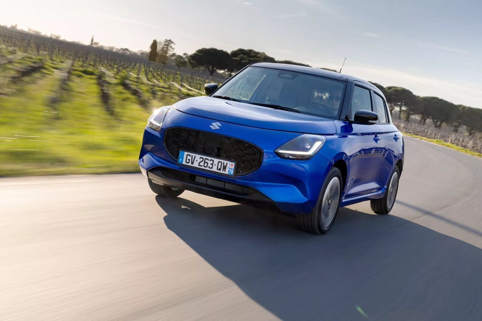 Modrý hatchback Suzuki Swift 7. generácie z roku 2024 idúci po zakrivenej asfaltovej ceste s LED svetlometmi, športovou prednou mriežkou, integrovanými hmlovými svetlami a aerodynamickým dizajnom viditeľným zo šikmého uhla spredu