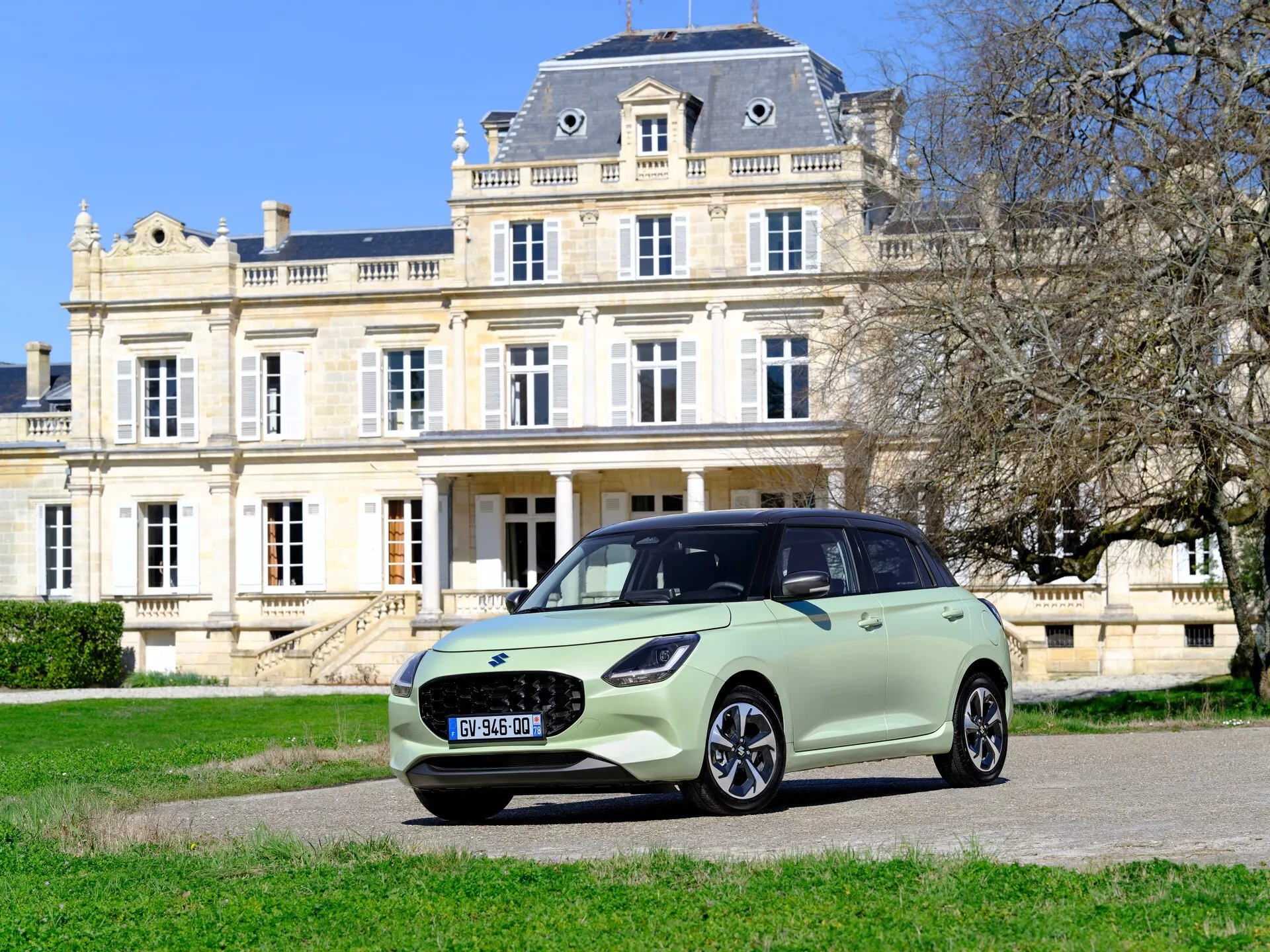 Hatchback Suzuki Swift 7. generácie, ročník 2024, zaparkovaný pred veľkolepým sídlom za jasného dňa, s LED svetlometmi, elegantnou prednou mriežkou, aerodynamickým dizajnom s dvojfarebným lakovaním a zliatinovými kolesami.