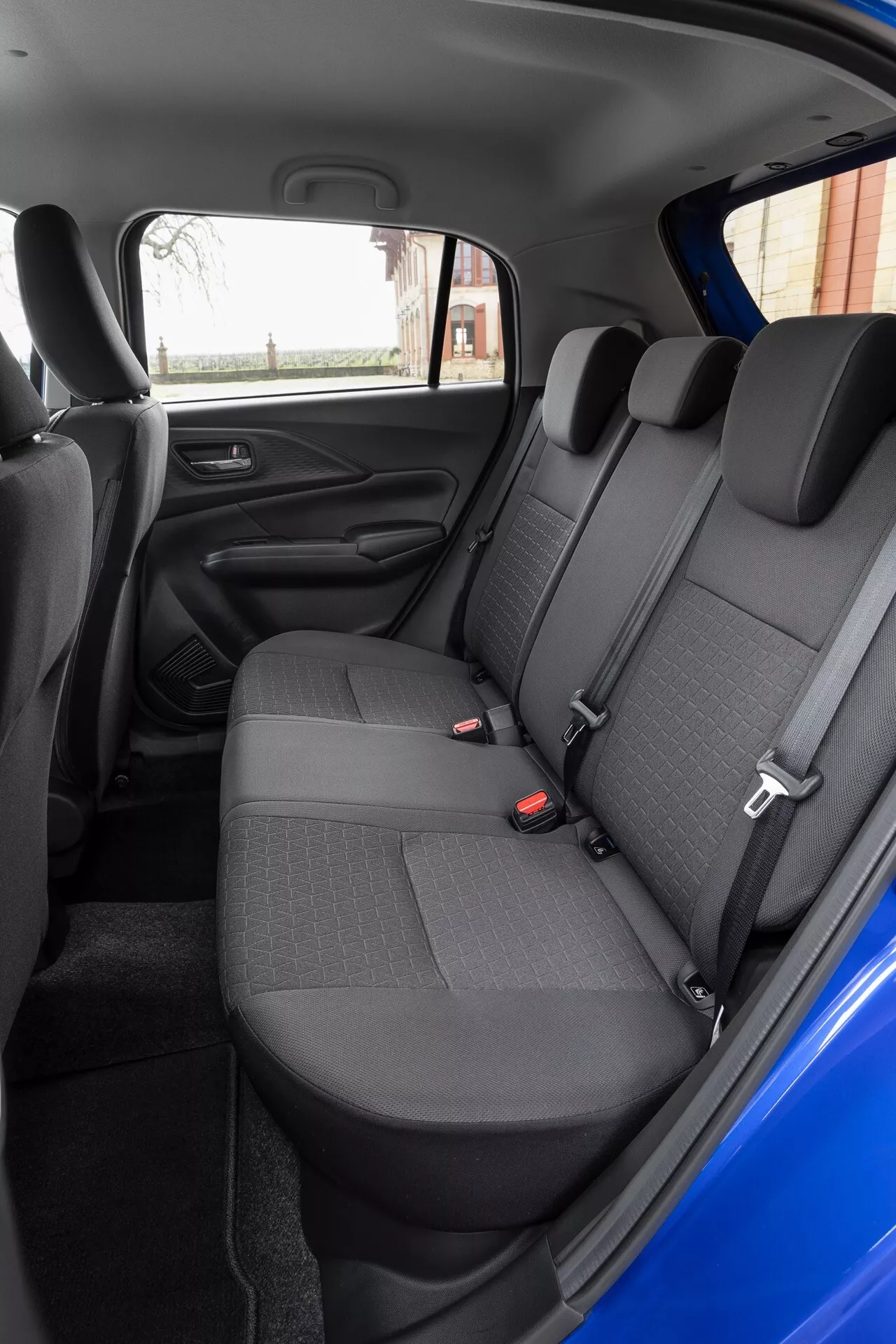 Interiér zadnej časti sedadiel Suzuki Swift 7. generácie 2024 s jasným výhľadom z pravej strany, s tmavošedými sedadlami z látky s dizajnom vzoru, funkciou deleného skladania, štandardnými bezpečnostnými pásami a elektrickým ovládaním okien na paneli zadnej bočnej dverí