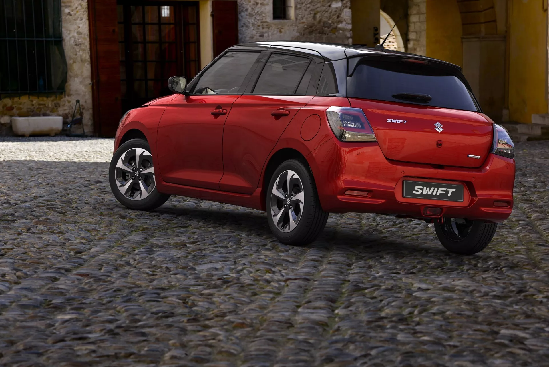 Červený hatchback Suzuki Swift 7. generácia 2024, pohľad z trojštvrťovej zadnej perspektívy ukazujúci zadný nárazník, veko batožinového priestoru, časť strechy, zadné čelné sklo, súpravu LED zadných svetiel, bočnú stranu cestujúceho, zliatinové kolesá, viditeľné značenie a anténu v tvare žraločej plutvy, zaparkovaný na dlažobných kockách.