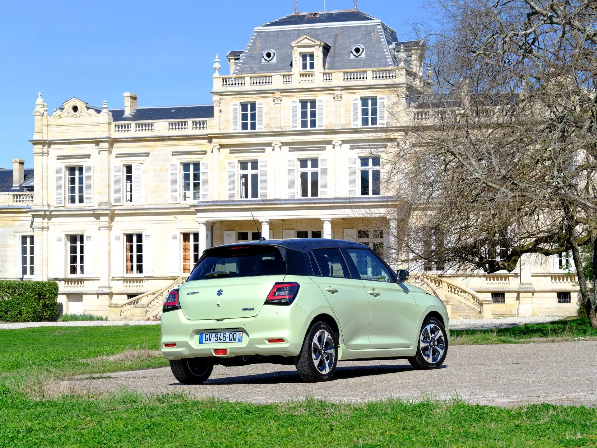 Suzuki Swift 7. generácia roku 2024 v bledo zelenej farbe, zobrazenie z uhla zozadu a zo strany s LED zadnými svetlami, zliatinovými kolesami a parkované na tráve oproti luxusnému sídlu na pozadí
