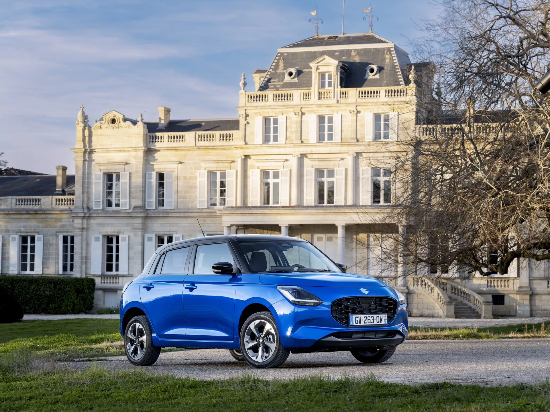 Modré kompaktné SUV Suzuki Swift 7. generácie z roku 2024 zaparkované na štrku pred klasickou budovou, predvádzajúce dizajnové prvky ako sú LED svetlomety, mriežka, zliatinové kolesá a elegantná silueta