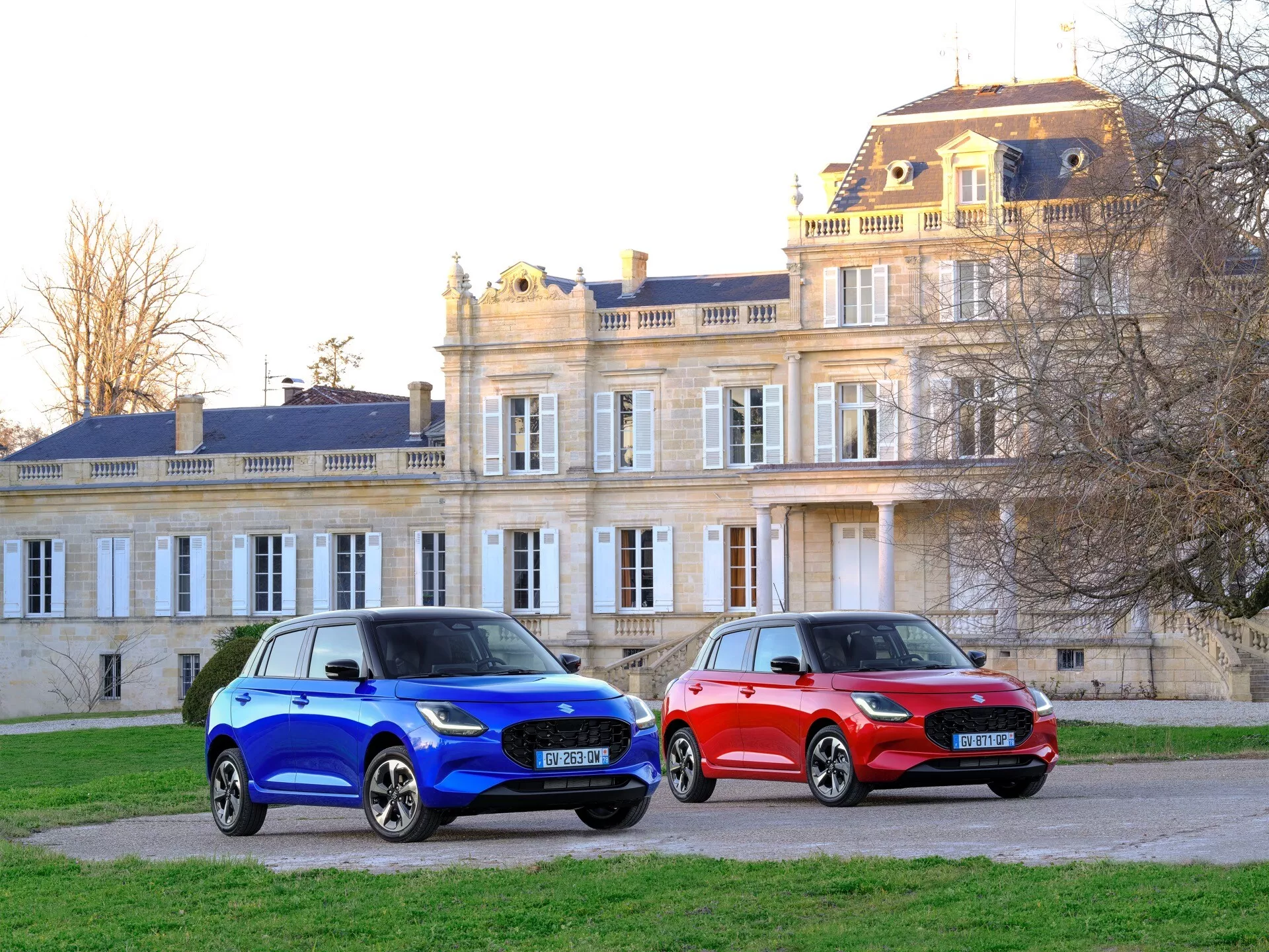 Dve autá Suzuki Swift 7. generácie ročníka 2024 zaparkované na štrku pri veľkolepom budove, predvádzajúce metalickú modrú a jasnú červenú farbu, moderné LED svetlomety, aerodynamické mriežky a disky z ľahkých zliatin v mäkkom svetle rána alebo neskorého popoludnia