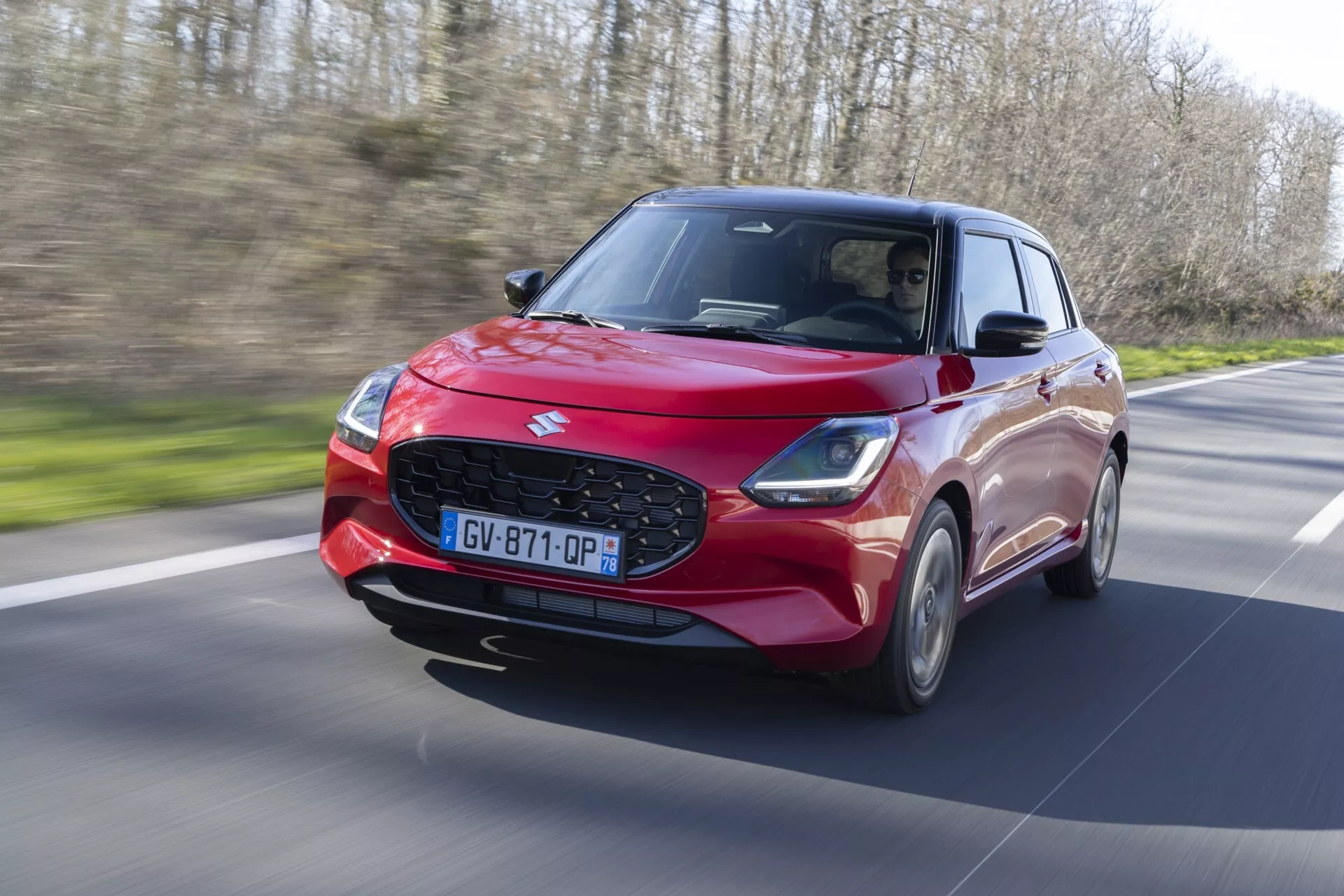 Suzuki Swift 2024, 7. generácia, v pohybe na vidieckej ceste, ukazujúci dynamický dizajn červeného hatchbacku, LED svetlomety a pokročilú technológiu asistencie vodiča vo svetlom dennom prostredí