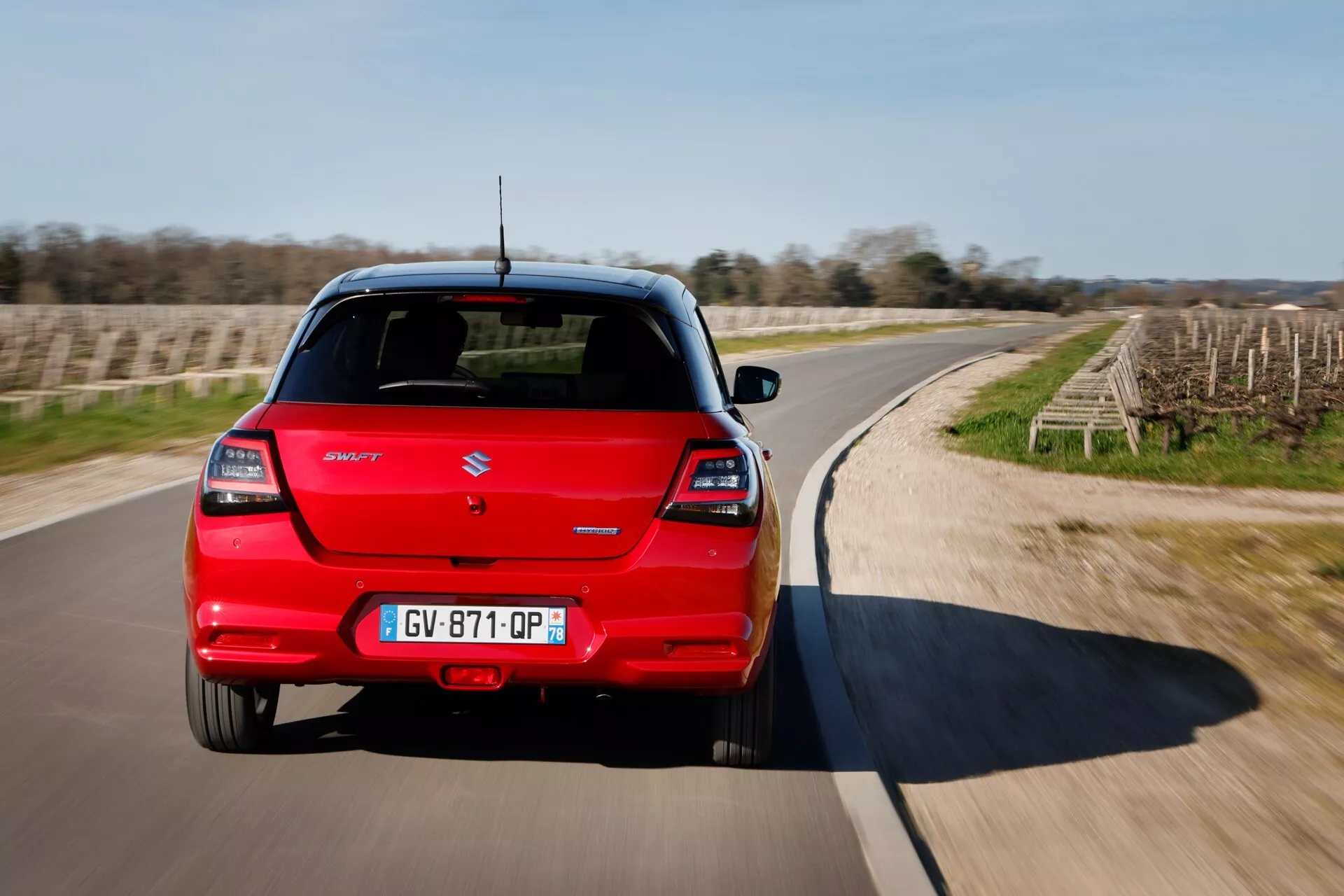 Suzuki Swift siedmej generácie 2024 v červenej farbe, zadný pohľad ukazuje batožinový priestor, LED svetlá a dynamický pohyb na zakrivenom ceste pod jasným nebom