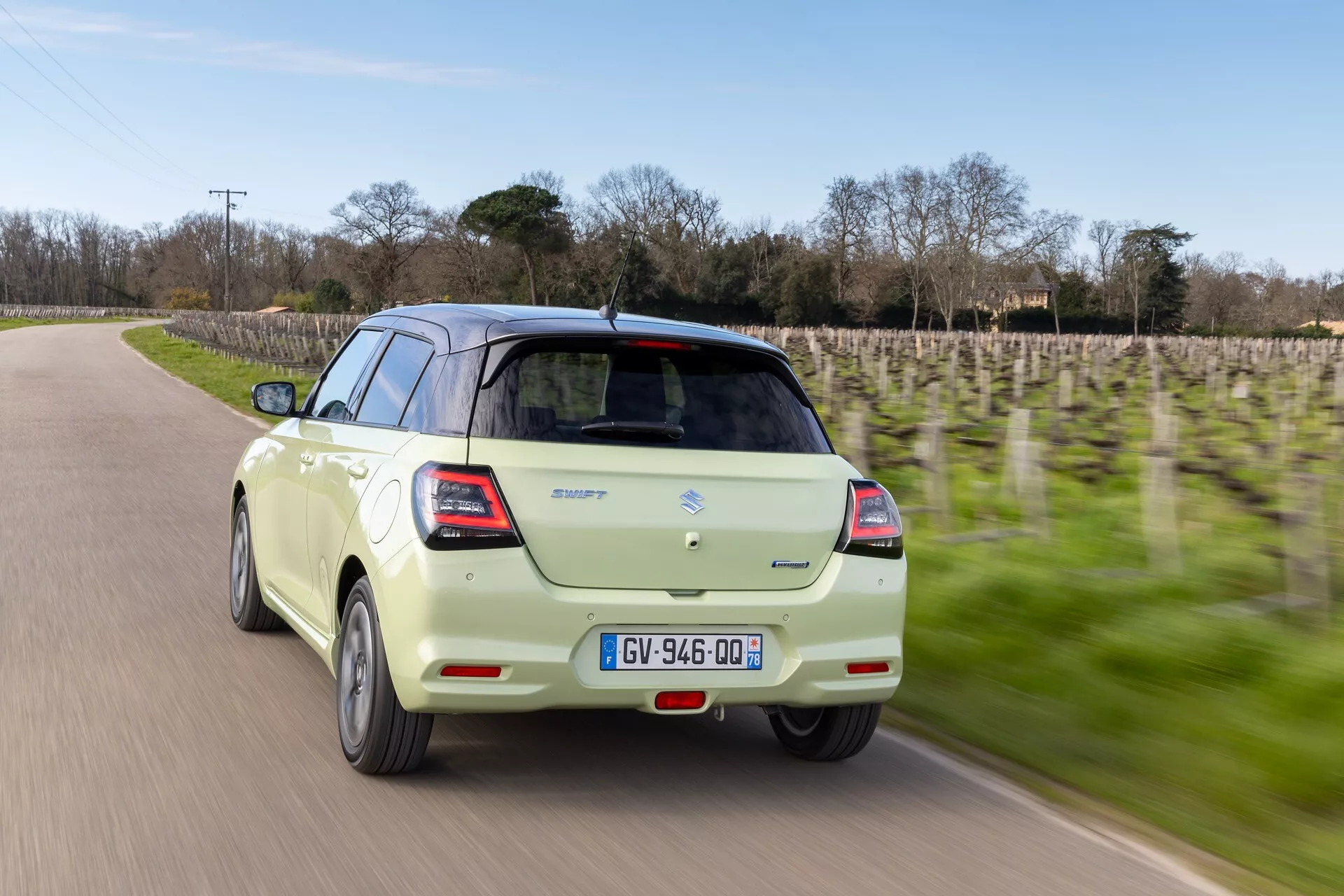 Suzuki Swift 7. generácie 2024 v pohybe, zachytené z pohľadu troch štvrtín zozadu so zobrazením kufra, zadného nárazníka, časti zadného čelného skla a bočných panelov na pozadí rozostreného vinohradu indikujúceho rýchlosť