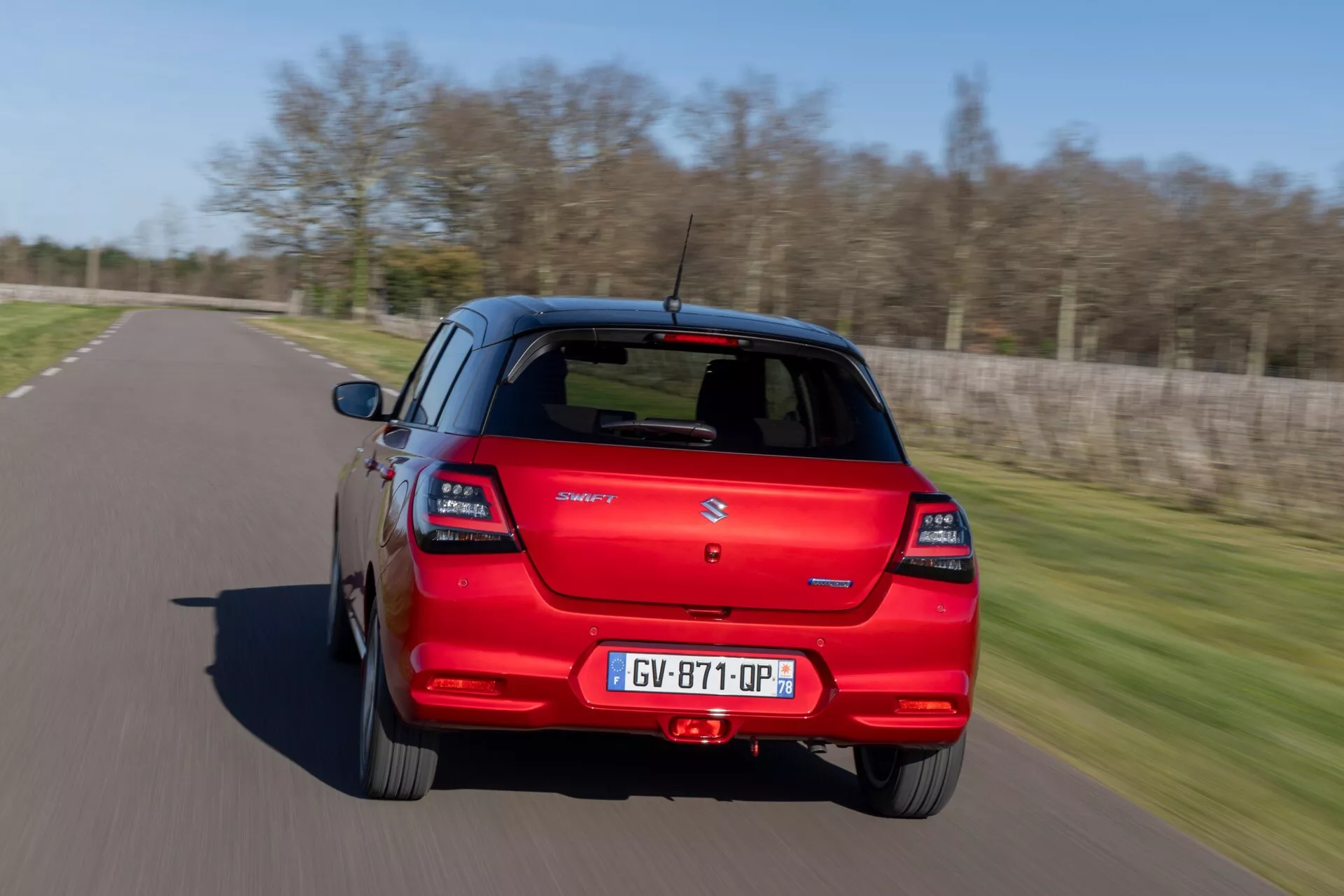 Suzuki Swift 7. generácie 2024 v červenej farbe ukazujúci zadný a bočný profil s moderným dizajnom hatchbacku, zadnými LED svetlami a rozostrením pohybu naznačujúcim pohyb, zachytené z mierne zvýšeného uhla