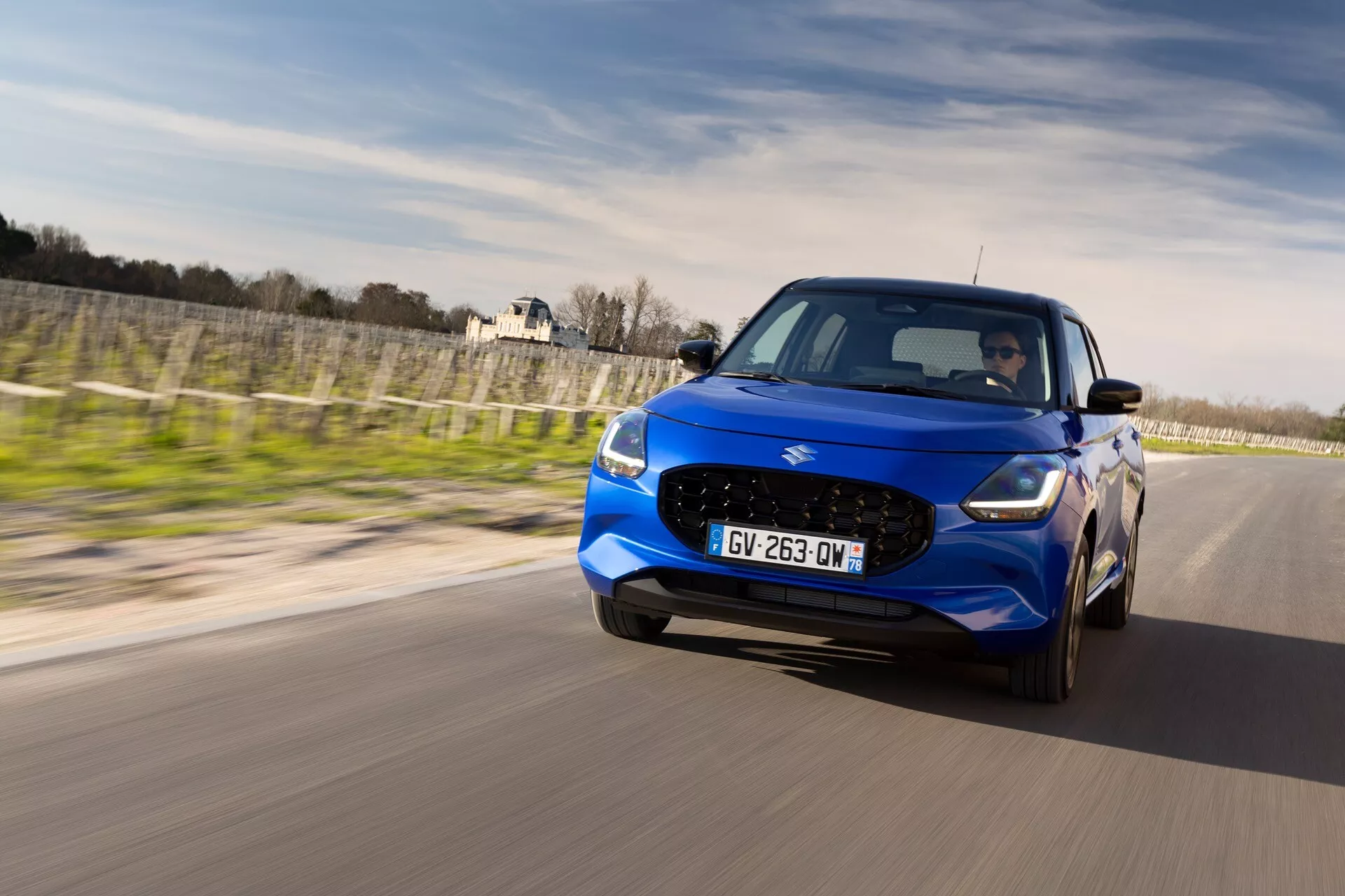 Suzuki Swift 7. generácie 2024 vo vibrantnej modrej, prezentujúci pokročilé dizajnové prvky vrátane odlišnej mriežky, štýlových LED svetlometov a športových kolies, zachytených v dynamickom dennom akčnom zábere na asfaltovej ceste