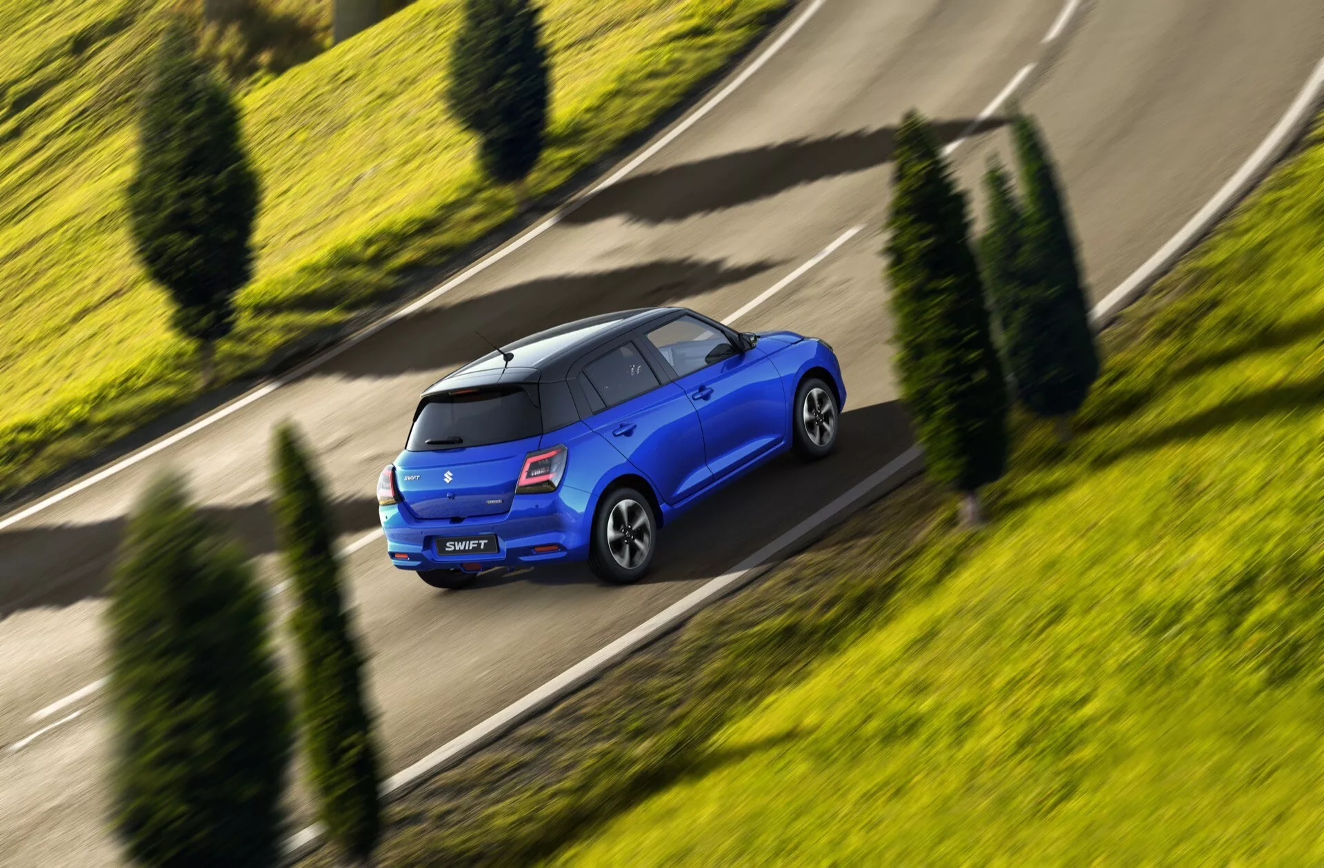 Suzuki Swift 2024, 7. generácia, modrý hatchback zachytený z vysokého uhla, ukazujúci jeho dynamický pohľad zozadu a z boku v pohybe na zakrivenom cieste, zdôrazňujúci strešný spojler, obopínajúce zadné svetlá a disky z ľahkej zliatiny v zelenom mestskom prostredí