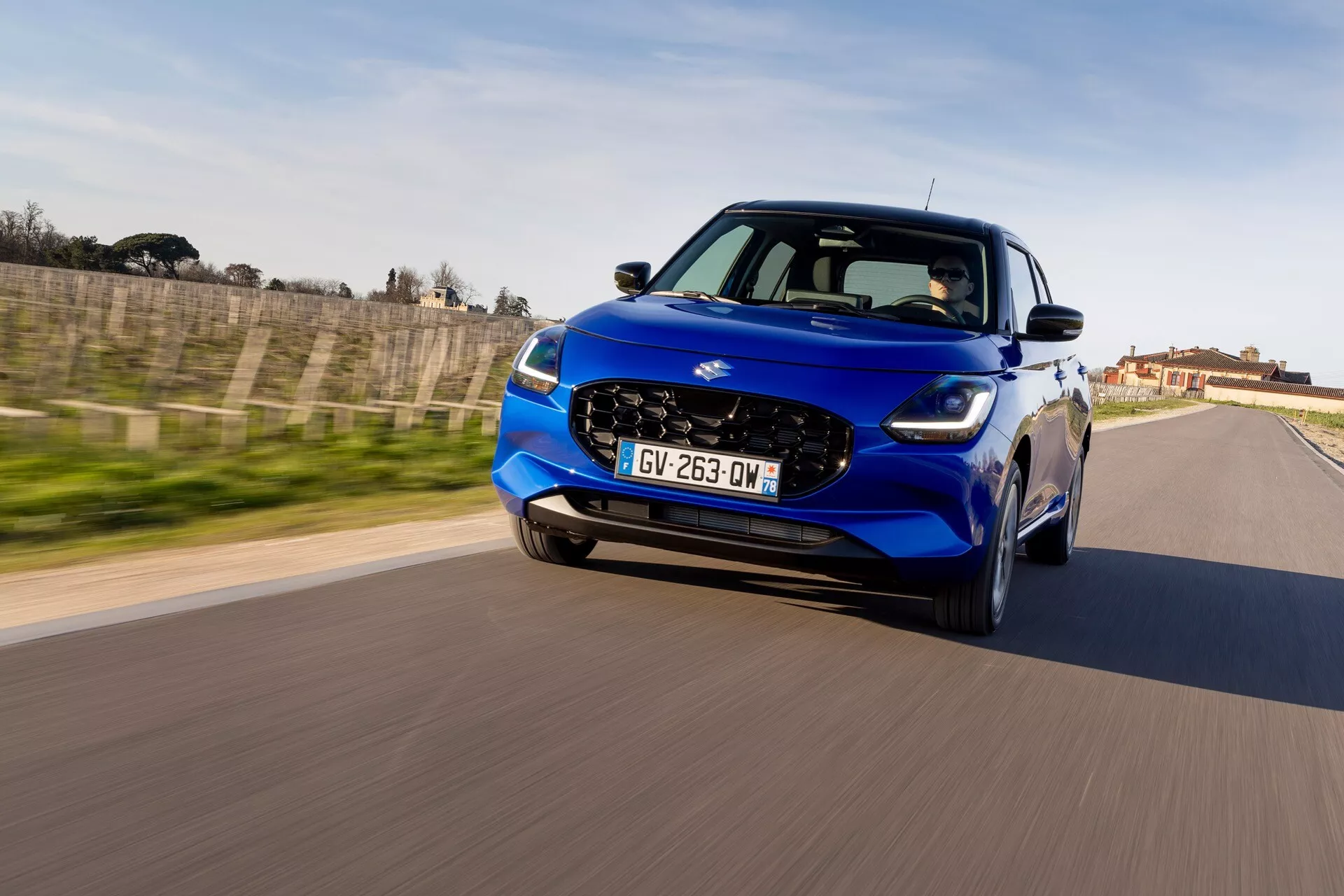 Suzuki Swift 2024 (7. generácia) v dynamickom, z nízkeho uhla, z pohľadu tri štvrtiny zpredu, predvádzajúci LED svetlomety, šesťuholníkovú mriežku a aerodynamický dizajn v pohybe na ceste
