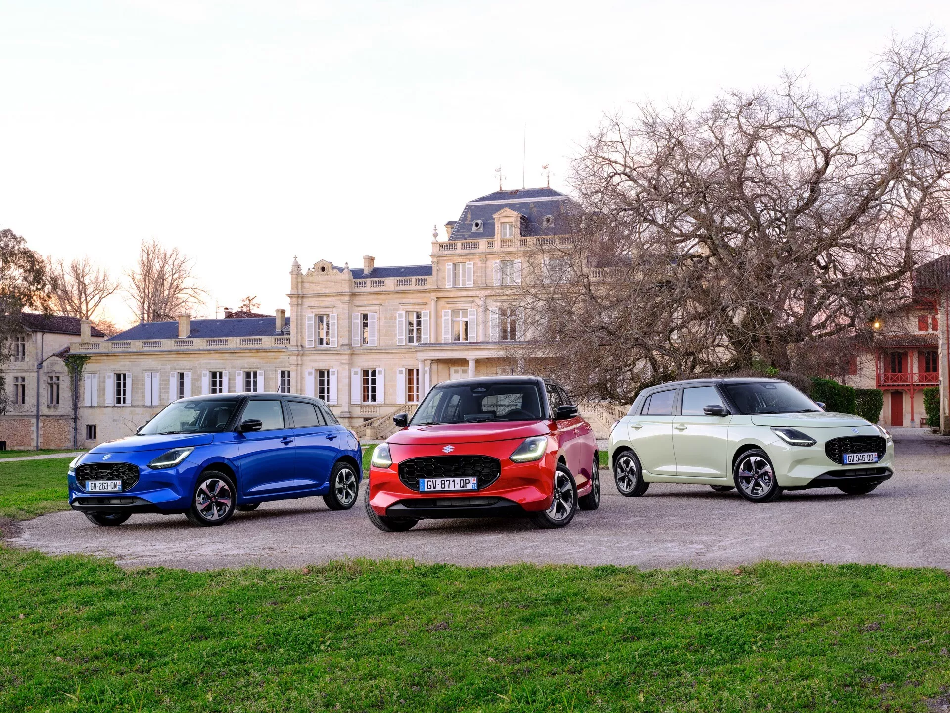 Suzuki Swift 2024, 7. generácia, s tromi modelmi vystavenými vedľa seba na pozadí klasickej architektúry, zdôrazňujúci moderný dizajn hatchbackov, pokročilú technológiu osvetlenia a aerodynamické prvky z čiastočnej prednej a bočnej perspektívy