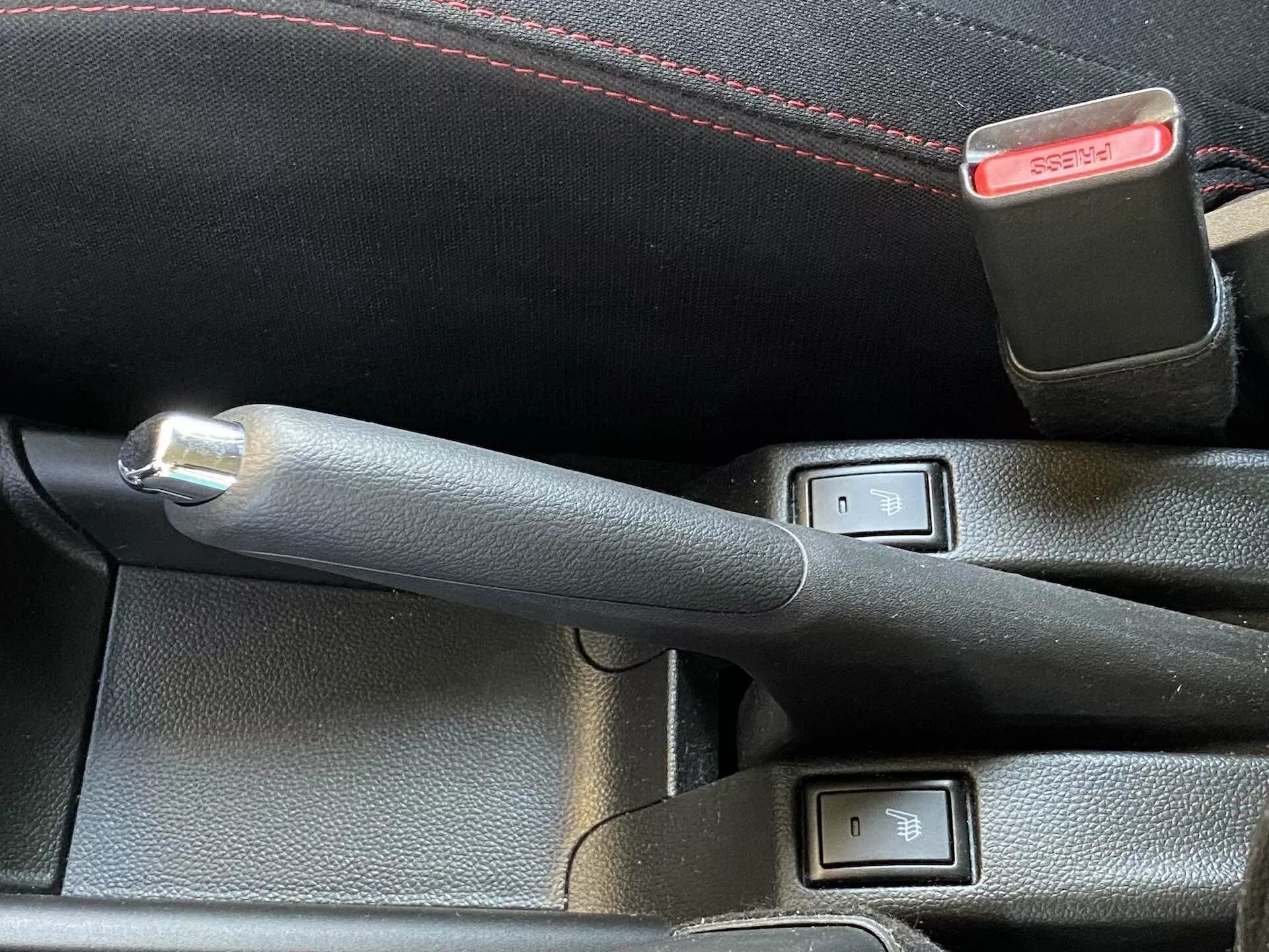Detailný pohľad na interiér Suzuki Swift 2017 (Swift Sport) zameraný na koženú ručnú brzdu s kovovým tlačidlom a ovládacími prvkami vyhrievania sedadiel. Ručná brzda je v strede fotografie a mierne naklonená doľava, čo naznačuje, že je zapnutá. Dve tlačidlá s LED indikátormi a ikonou sedadla vedľa ručnej brzdy naznačujú dvojzónové vyhrievanie predných sedadiel. Za ovládacími prvkami je čiastočne viditeľná spona bezpečnostného pásu s červeným uvoľňovacím tlačidlom. Obrázok je urobený z vyvýšeného uhla pri dennom svetle, zdôrazňujúc technológické a pohodlne prvky.