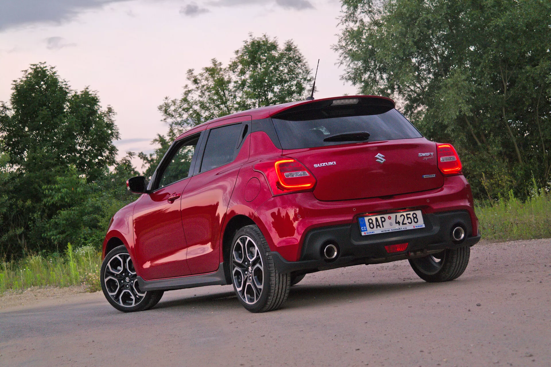 Červený Suzuki Swift Sport 2017 hatchback zachytený zozadu z troch štvrtinového uhla, zdôrazňujúci jeho zadný a bočný profil na strane vodiča. Fotografia, urobená z mierne zníženej perspektívy, zdôrazňuje moderné viacramenné hliníkové kolesá a nízkoprofilové pneumatiky. Svetelné podmienky pri svitaní alebo súmraku s občasnou oblačnosťou poskytujú mäkké prirodzené svetlo. Viditeľné funkcie zahŕňajú moderný dizajn zadných svetiel s LED prvkami, značku výrobcu, názov modelu, bočné zrkadlá vo farbe karosérie, ktoré môžu obsahovať smerové svetlá, zadný nárazník s integrovanými parkovacími senzormi a strešný spoiler pre aerodynamickú účinnosť.