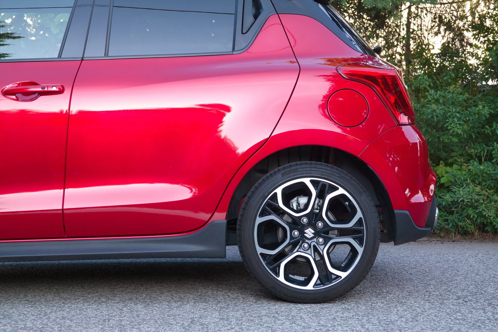 Čiastočný bočný pohľad na červený Suzuki Swift 2017 (Swift Sport), sústredený na zadnú časť vrátane zadného kolesa a oblasti blatníka. Detailný záber zachytáva štýlový zliatinový disk s viaclúčovým dizajnom, nízkoprofilovú pneumatiku, moderný zakrivený zadný blatník, hlboký lesklý červený lak, časť zadného svetla a chrómové ozdobné prvky, ktoré zvýrazňujú jeho športový dizajn a prémiovú povrchovú úpravu.