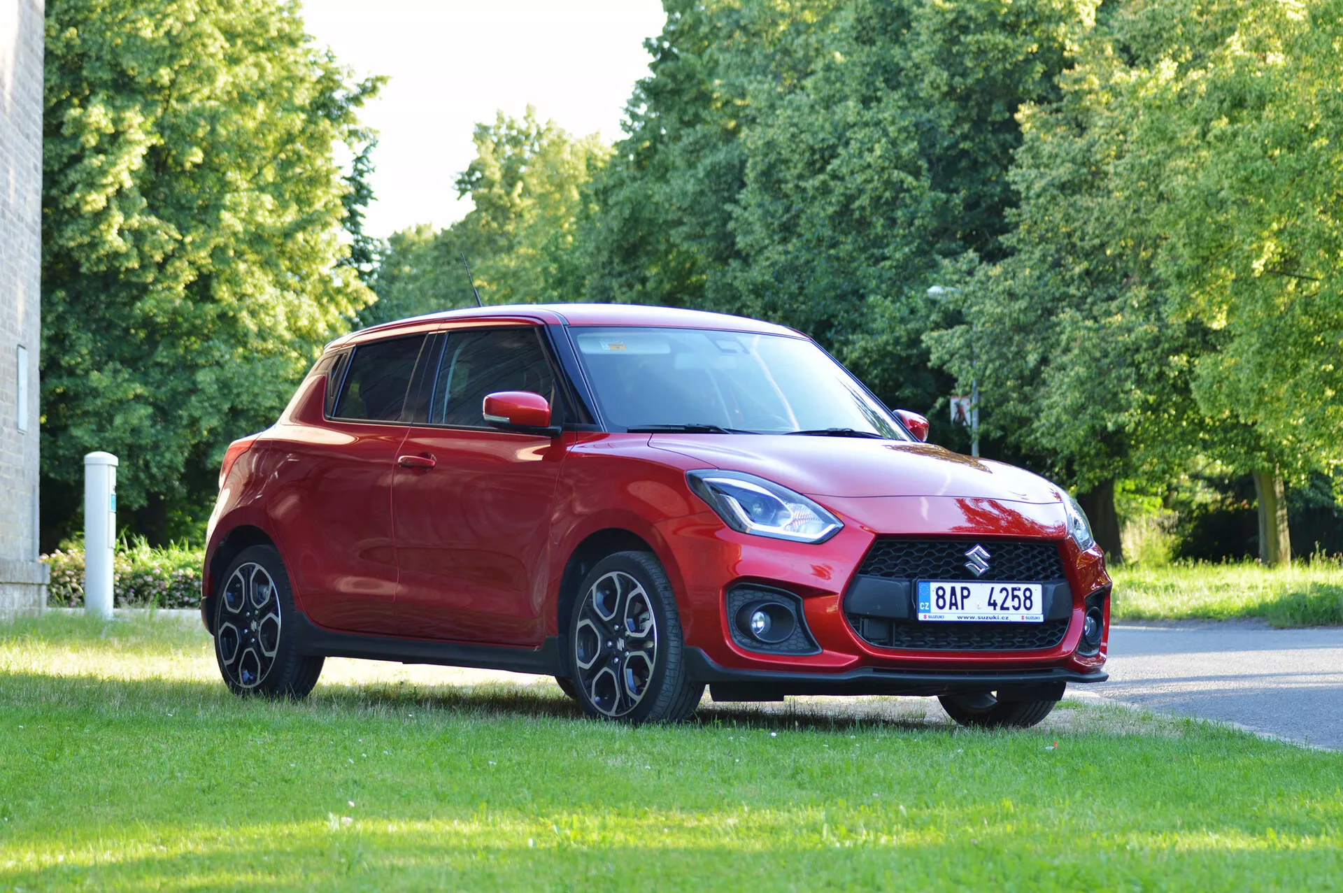Obraz zobrazuje červený hatchback Suzuki Swift Sport 2017 umiestnený na trávnatom povrchu, pravdepodobne v parku alebo pri ceste, zachytený z predného trojštvrťového uhlu. Výrazná predná mriežka, elegantné a uhlové svetlomety, zostava hmloviek a sklonená kapota do aerodynamického čelného skla vynikajú. Zliatinové kolesá, bočné zrkadlá v farbe karosérie, jemne sa zvažujúca línia strechy k zadnému oknu a tónované okná prispievajú k dizajnu vozidla. Prírodné osvetlenie a neblokovaný výhľad zdôrazňujú celkový postoj a dizajn auta.