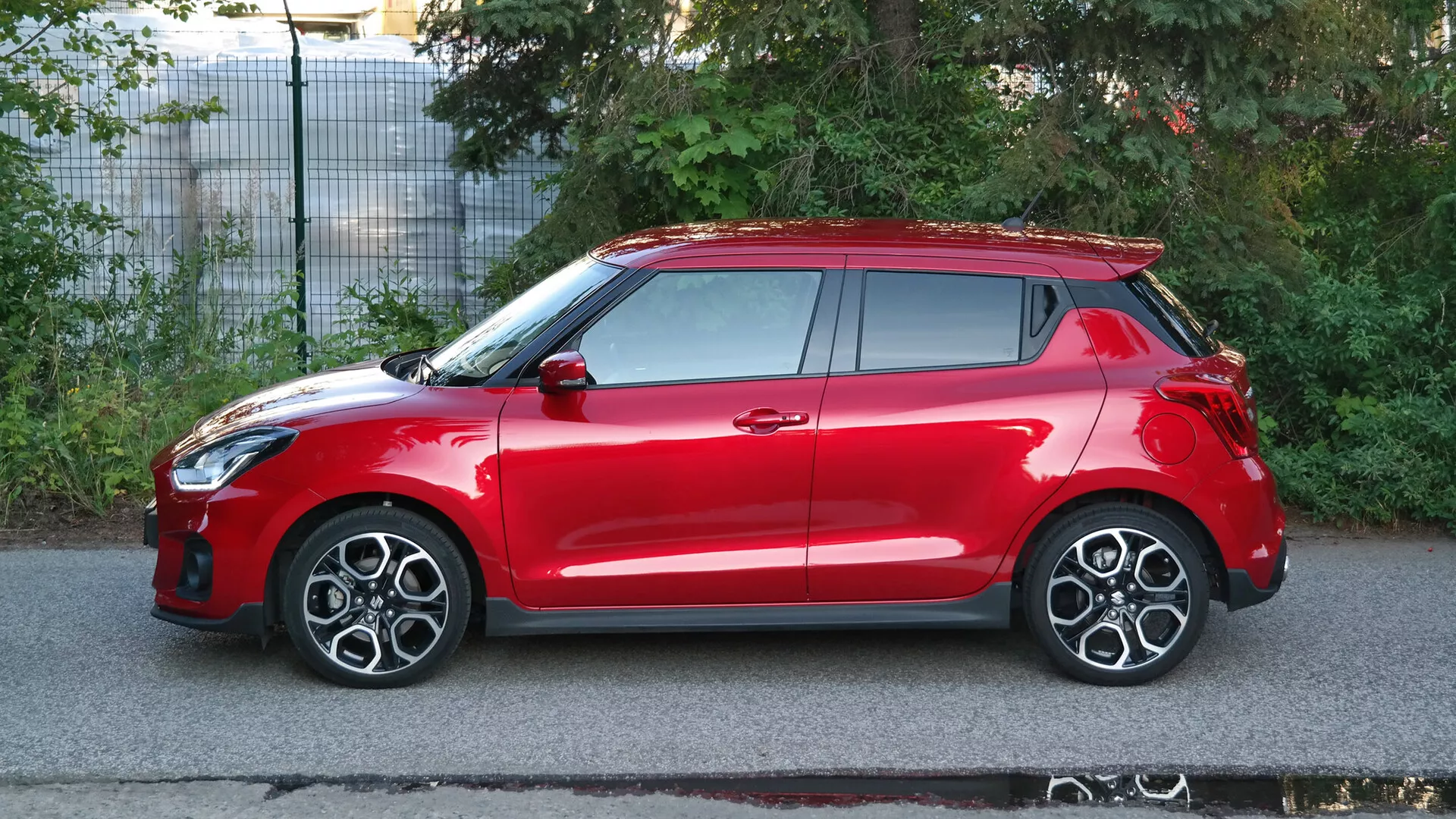 Bočný profilový pohľad na červený hatchback Suzuki Swift Sport z roku 2017. Vozidlo je zachytené za denného svetla, čo zvýrazňuje jeho elegantné dizajnové prvky vrátane integrácie svetlometov do predného oblúku kolesa, viacpaprskových zliatinových kolies, bočných zrkadiel a kľučky dverí vo farbe karosérie, systému bezkľúčového vstupu a viditeľného strešného okna. Auto tiež disponuje strešným spojlerom a vysoko umiestnenými zadnými svetlami. Obrázok, urobený na predmestí, zdôrazňuje aerodynamiku vozidla, jeho športové charakteristiky a pravdepodobne pokročilé bezpečnostné technológie.
