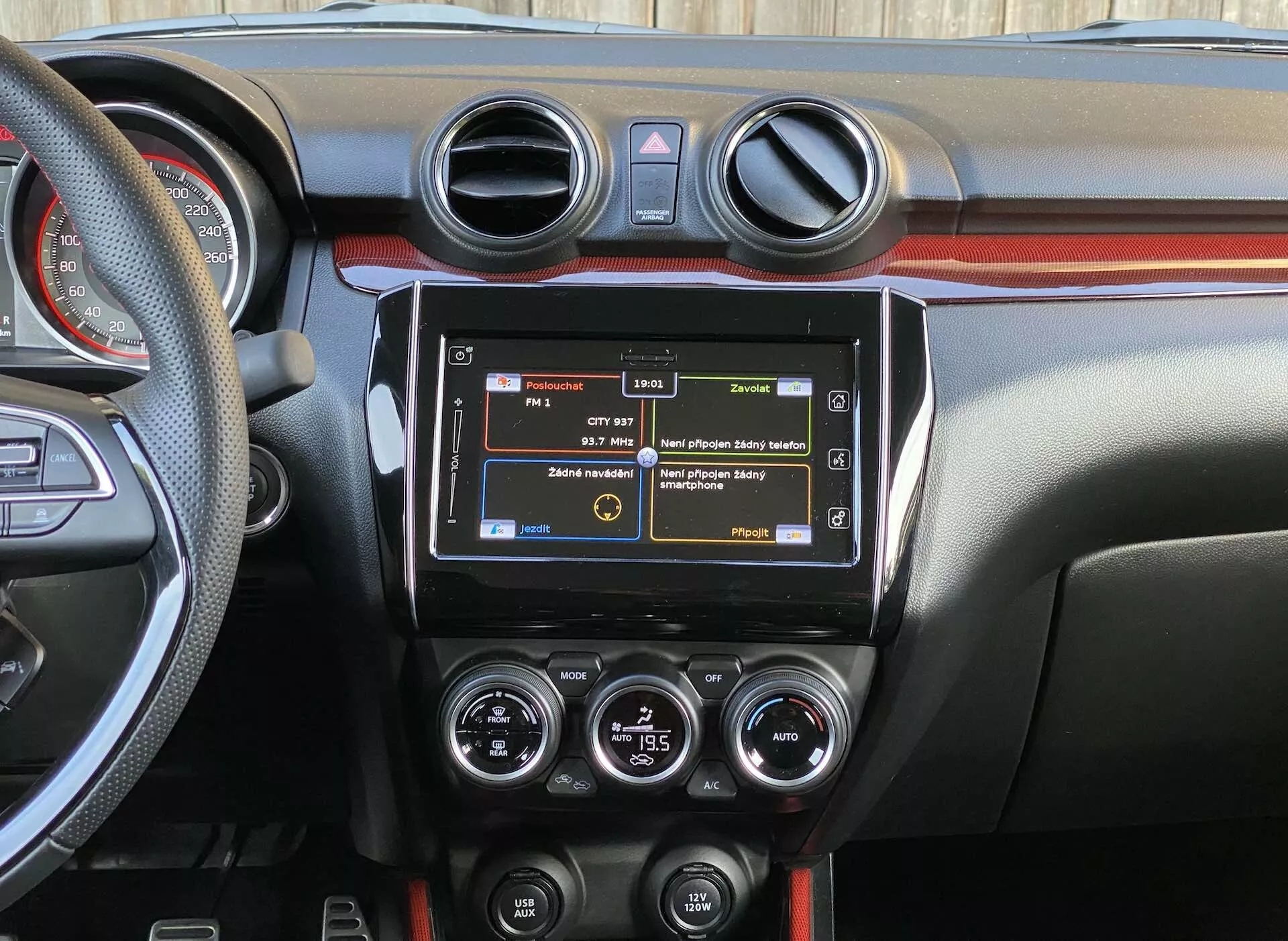 Detailný pohľad na interiér Suzuki Swift Sport 2017 zameraný na stredový palubný panel z pohľadu zadného sedadla, zobrazujúci moderný infotainment systém, kruhové ventilačné otvory klimatizácie, ovládacie prvky HVAC, rôzne funkčné tlačidlá, dvojfarebný dizajn palubnej dosky a časť volantu s namontovanými ovládačmi.