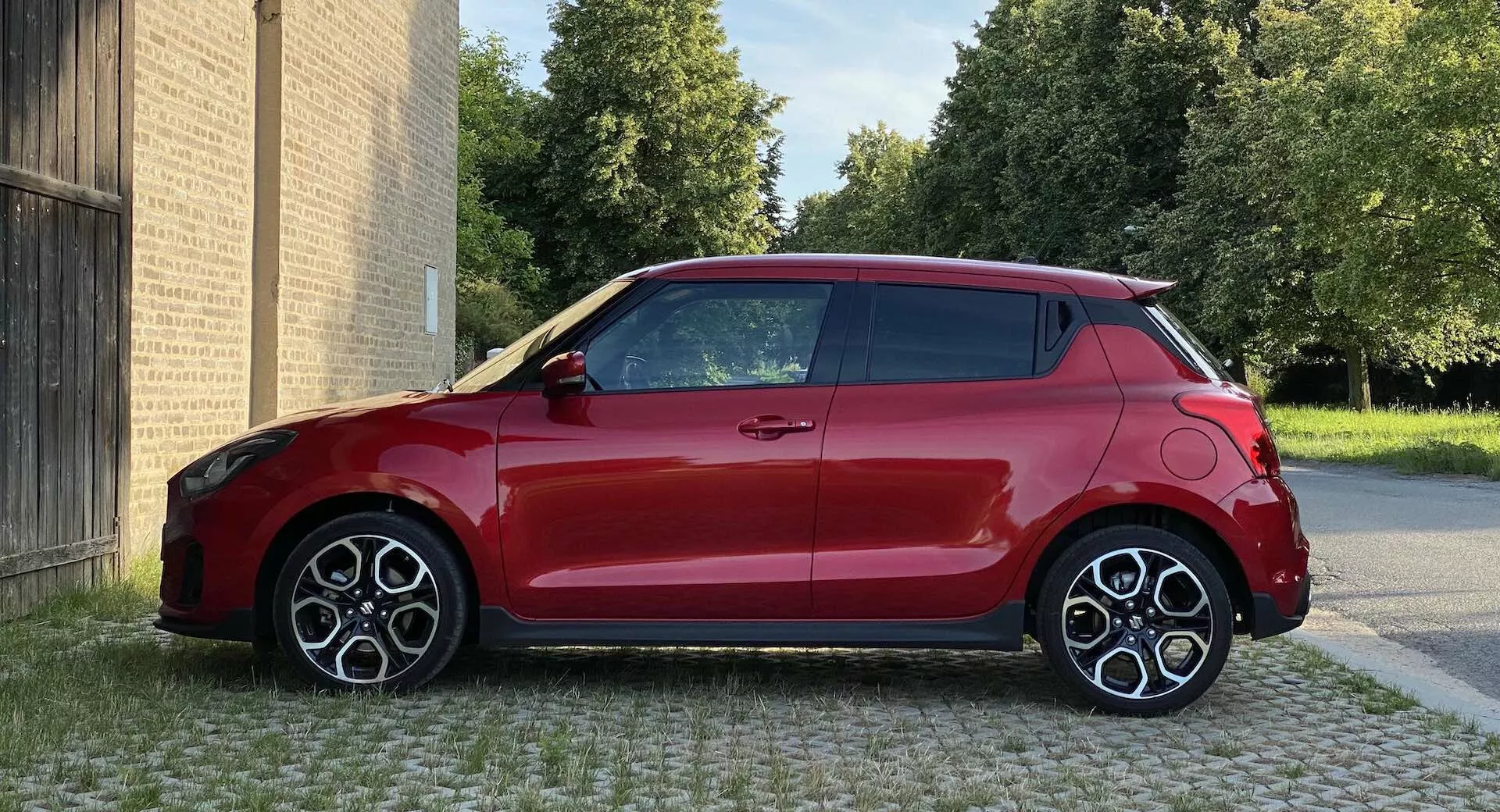 Obrázok červeného hatchbacku Suzuki Swift Sport 2017, zhotovený z bočného profilu na úrovni očí, zobrazujúci pravú stranu s dvojtónovým dizajnom červeného karosérie a čiernej strechy. Auto je zaparkované na dláždenej ploche s budovami v pozadí a stromami viditeľnými za budovami. Kompaktný, moderný dizajn má veľké športové kolesá s viacerými lúčmi, viditeľné aerodynamické línie a anténu v tvare plutvy žraloka. Osvetlenie je denné, kvalita obrazu je ostrá a viditeľné sú povrchové odrazy.