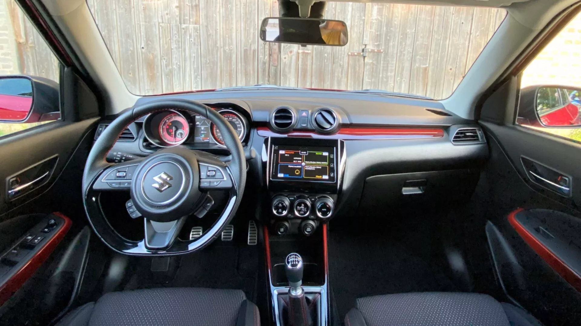 Pohľad na interiér Suzuki Swift 2017 (Swift Sport) z pohľadu zadného sedadla smerom k prednej kabíne, zahrňujúci detailné zobrazenie palubnej dosky, volantu s integrovanými ovládacími prvkami, centrálnej konzoly s infotainment systémom a ovládaním klimatizácie, automatickú prevodovku a čierne interiérové materiály s červenými akcentmi.