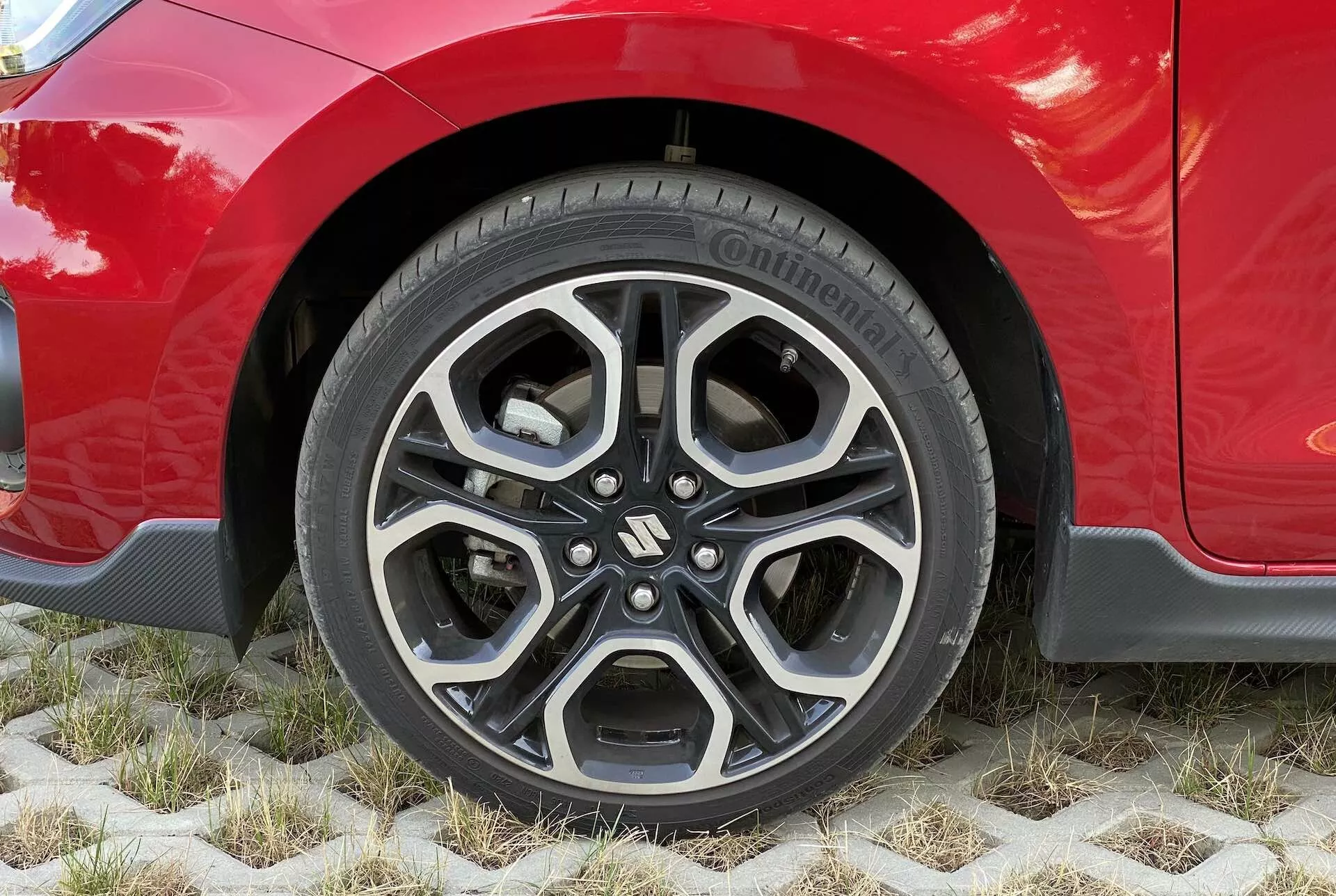 Zblízka na koleso červeného Suzuki Swift 2017 Swift Sport, zobrazujúce moderný hliníkový disk s komplexným, viacramenným dizajnom v čiernej a obrábanej striebornej farbe. Viditeľná pneumatika je model Continental PremiumContact, čo naznačuje vysokú kvalitu a dobrý stav pneumatiky. Karoséria auta je nad kolesom lesklá a dobre udržiavaná, zvýrazňujúca estetické línie blatníka a možno aj športovú výbavu. Vozidlo je zaparkované na ekologických šesťhranných dlaždiciach umožňujúcich rast trávy, zdôrazňujúcich udržateľné parkovacie riešenia.