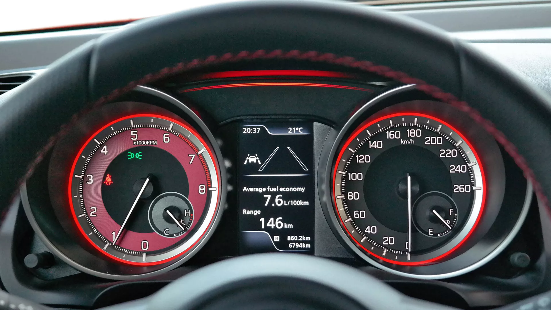 Detailný pohľad z prvej osoby na prístrojovú dosku vozu Suzuki Swift 2017 (Sport) ukazuje analógový otáčkomer (do 7 000 ot./min) a rýchlomer (do 260 km/h) s červenými akcentmi a stredový digitálny displej, ktorý zobrazuje čas 20:37, vonkajšiu teplotu 21°C, priemernú spotrebu paliva 7,6 l/100 km, zostávajúci dojazd 146 km, dva cestovné počítadlá (0,0 km a 367,6 km) a celkový počet najazdených kilometrov 80 622 km. Osvetlenie naznačuje, že vozidlo je v chode, pravdepodobne za obmedzených svetelných podmienok, čo odráža kombináciu analógovej a digitálnej technológie prístrojovej dosky.