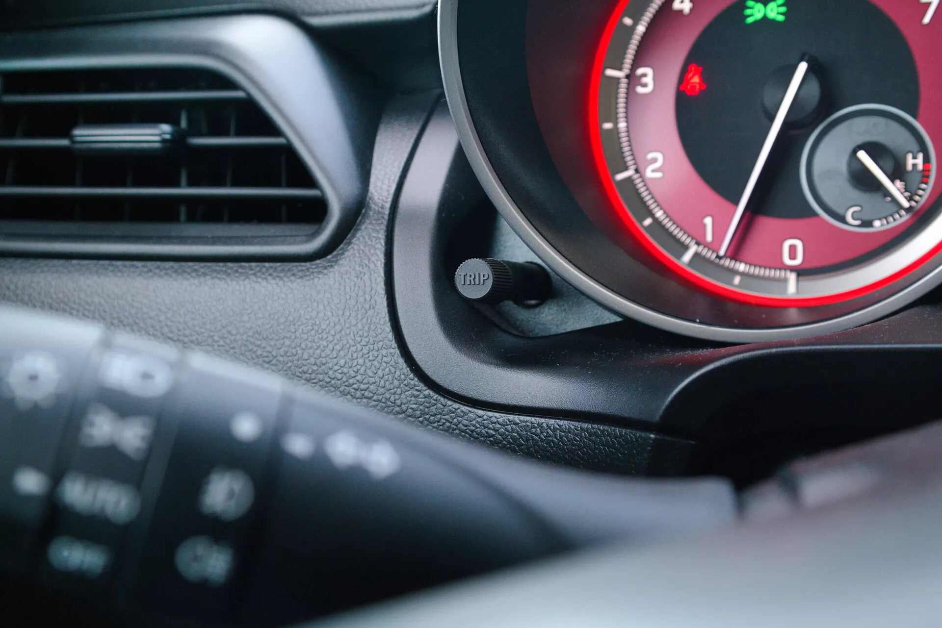 Interiér modelu Suzuki Swift Sport 2017 sa zameriava na prístrojový panel a časť volantu z pohľadu vodiča. Obrázok zvýrazňuje otáčkomer s červenými akcentmi, tlačidlo 'TRIP' a časť ďalšieho ukazovateľa, pravdepodobne teplotného. Multifunkčné ovládače na volante naznačujú integrované nastavenia audia a tempomatu.