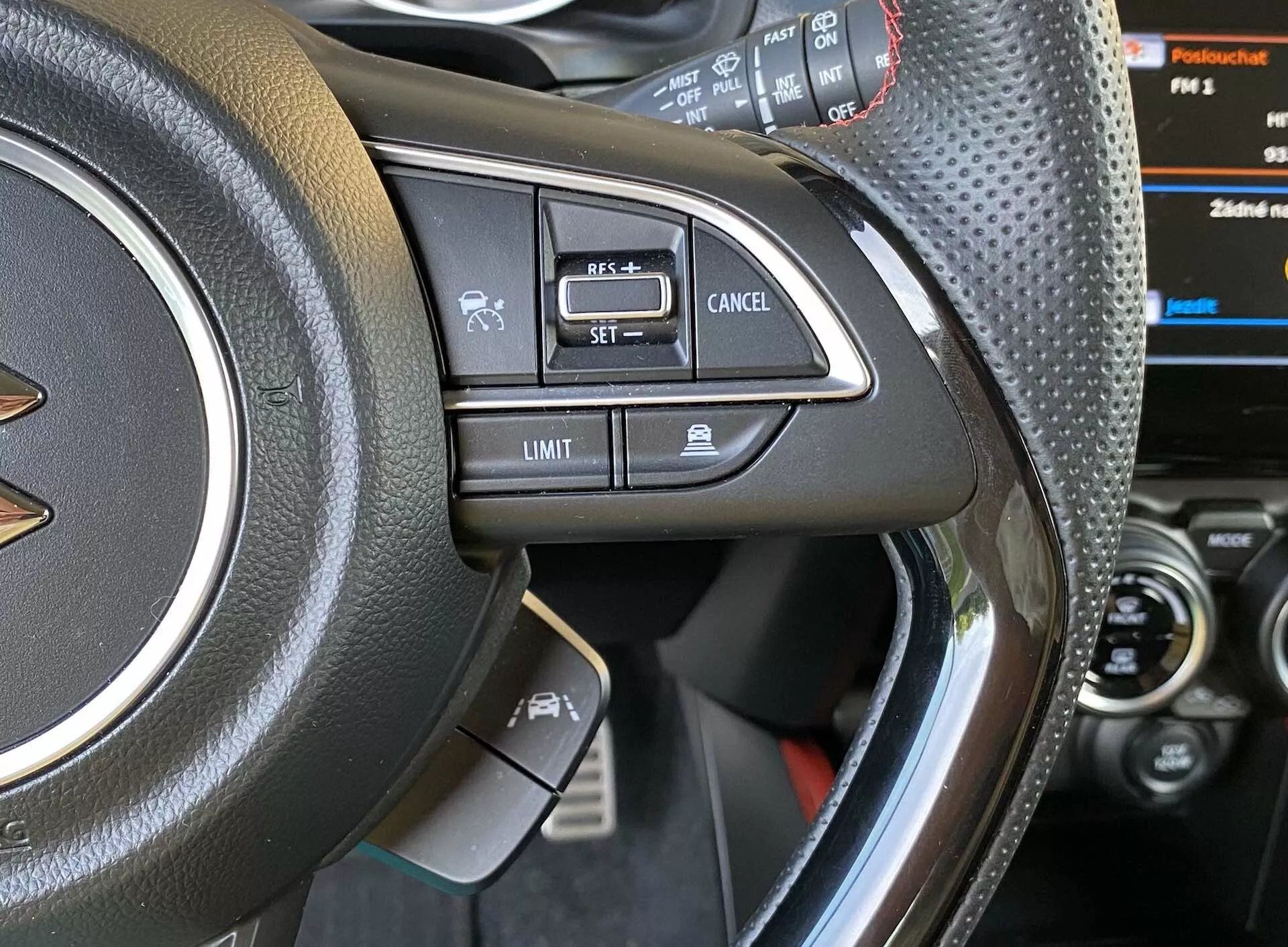 Detailný pohľad na ľavú stranu volantu Suzuki Swift 2017 Swift-Sport so zameraním na ovládacie prvky. Fotografia zhotovená zo sedadla vodiča alebo trochu mimo dverí vodiča, zameraná na volant z kože alebo koženky s prepravkou, kovový akcent a rôzne tlačidlá ako hlasové ovládanie, Bluetooth hands-free volanie, tempomat 'SET,' 'Cancel' a obmedzovač rýchlosti 'Limit.' Čiastočne viditeľný prístrojový panel s digitálnym displejom a stredovou konzolou.