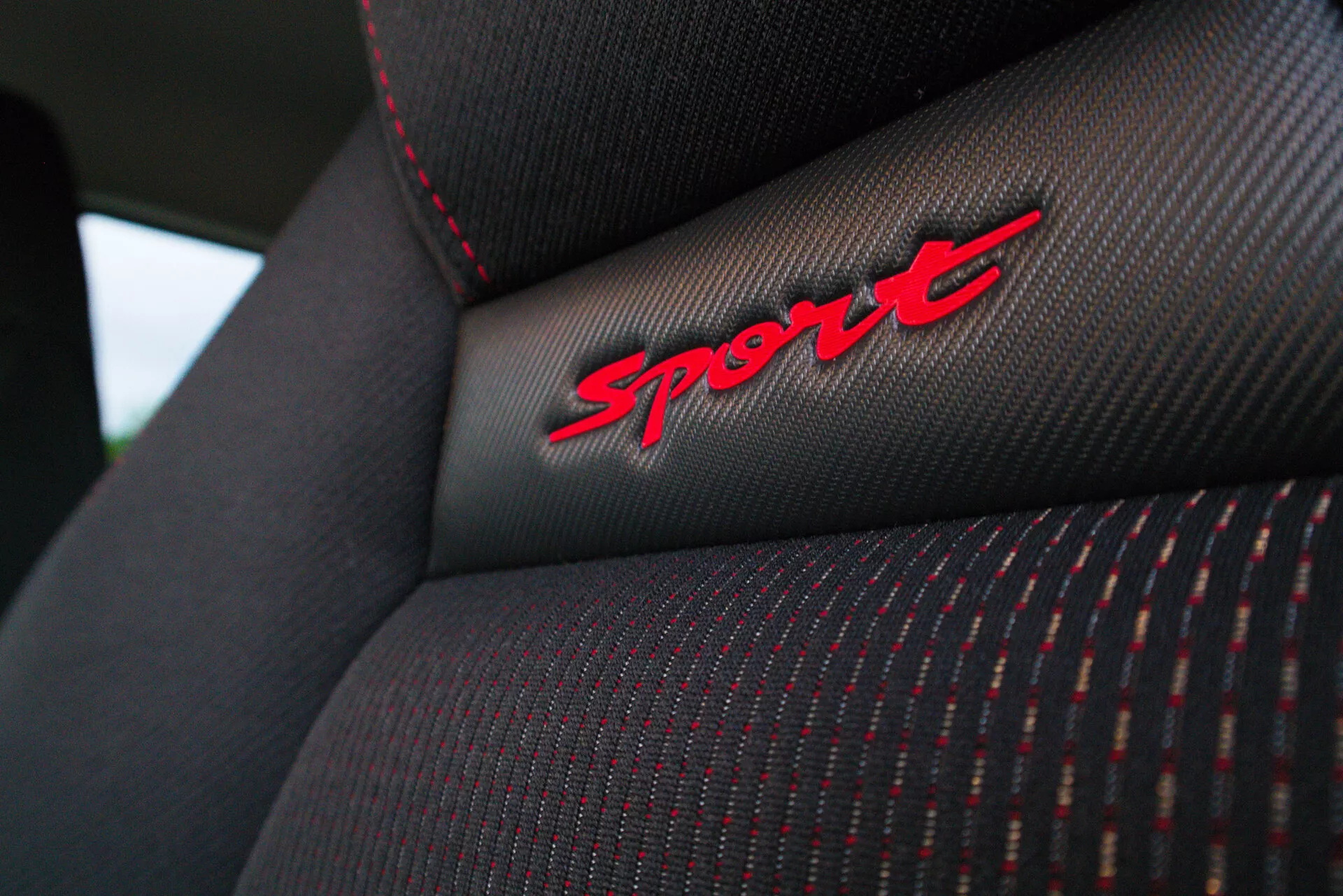 Detailný záber na operadlo sedadla Suzuki Swift Sport 2017, ktorý ukazuje textúru látky, červené švy a výšivku 'Sport' na čiernej látke, zdôrazňujúci športovú výbavu vozidla.
