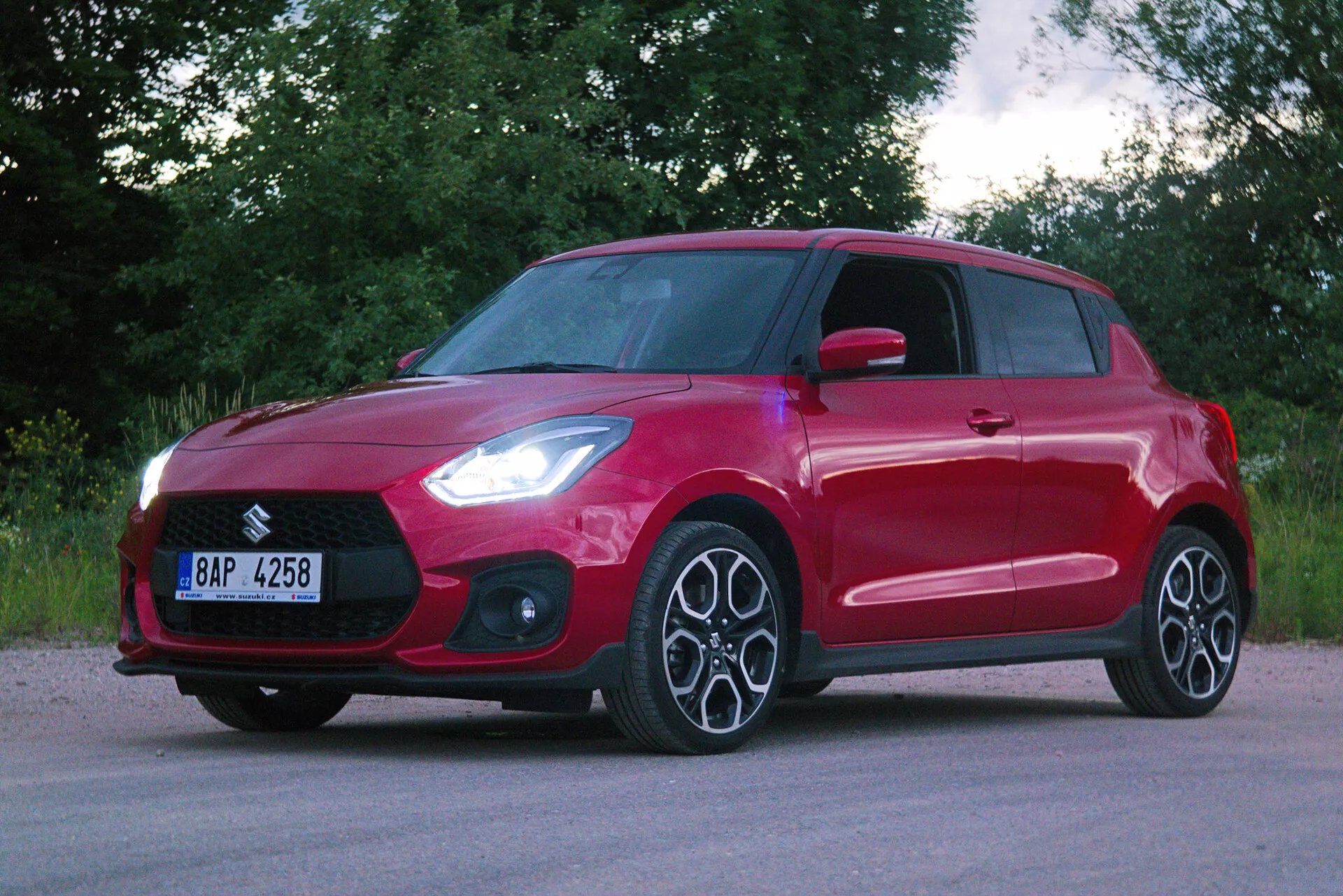 Trojštvrťový predný pohľad na červený hatchback, ktorý ukazuje moderné dizajnové prvky ako výraznú prednú mriežku, elegantné hranaté svetlomety pravdepodobne s LED technológiou, viaclúčové zliatinové disky a dynamickú kontúru línie na bočnom profile. Vozidlo má kľučky dverí v farbe karosérie a integrované smerové svetlá v spätných zrkadlách. Lesklý červený lak je rovnomerný a bez poškodenia, auto je zaparkované na asfaltovom povrchu s pozadím so zeleňou, čo naznačuje vidiecke alebo prímestské prostredie.