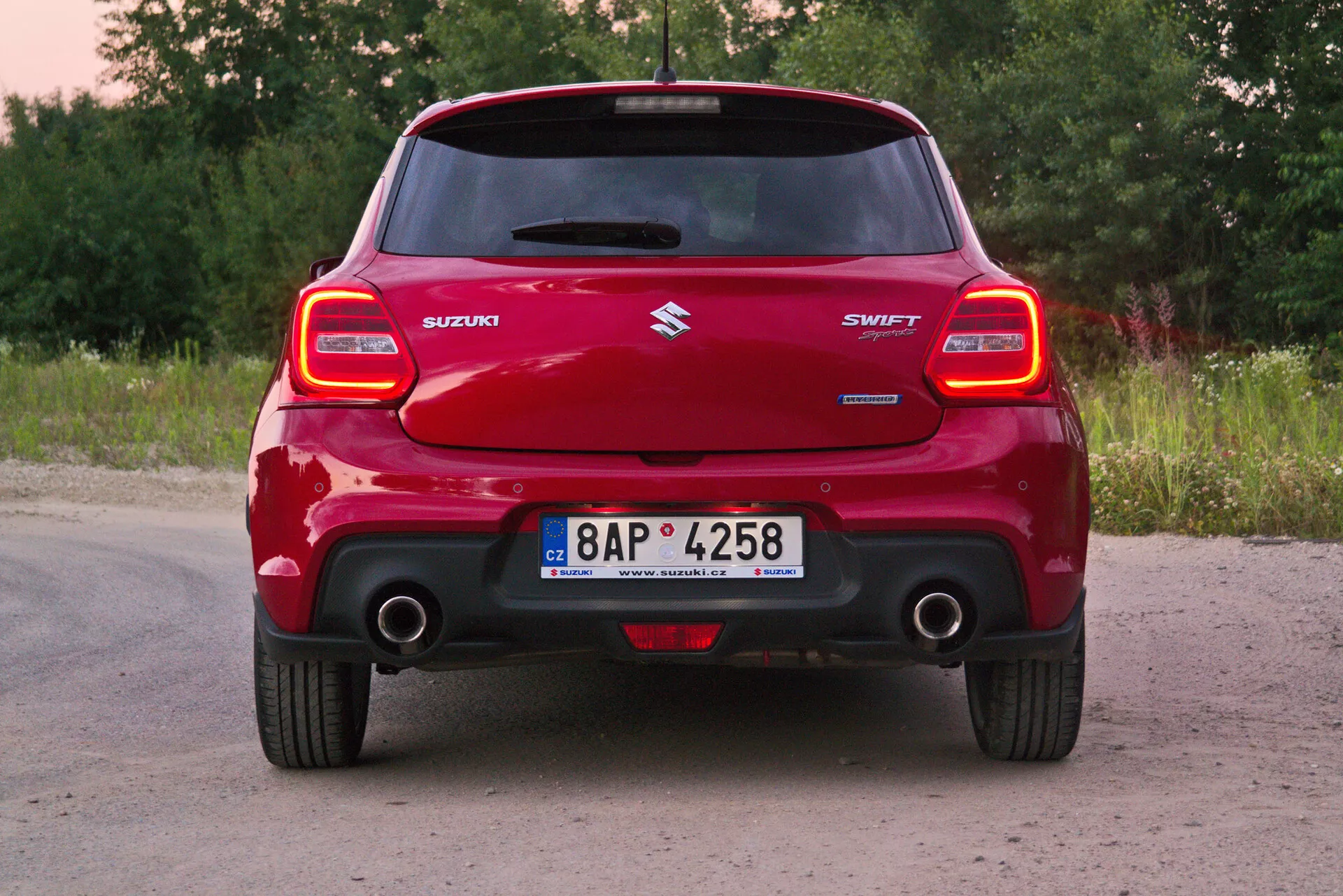 Zadný pohľad na červené auto Suzuki Swift Sport 2017, zameraný na celkovú šírku a výrazný zadný nárazník, charakteristické zadné svetelné zhluky, strešný spojler, centrálny logo Suzuki, názov modelu 'SWIFT', viditeľnú výfukovú trubku a registračnú značku v vidieckom prostredí so zeleňou a stromami.