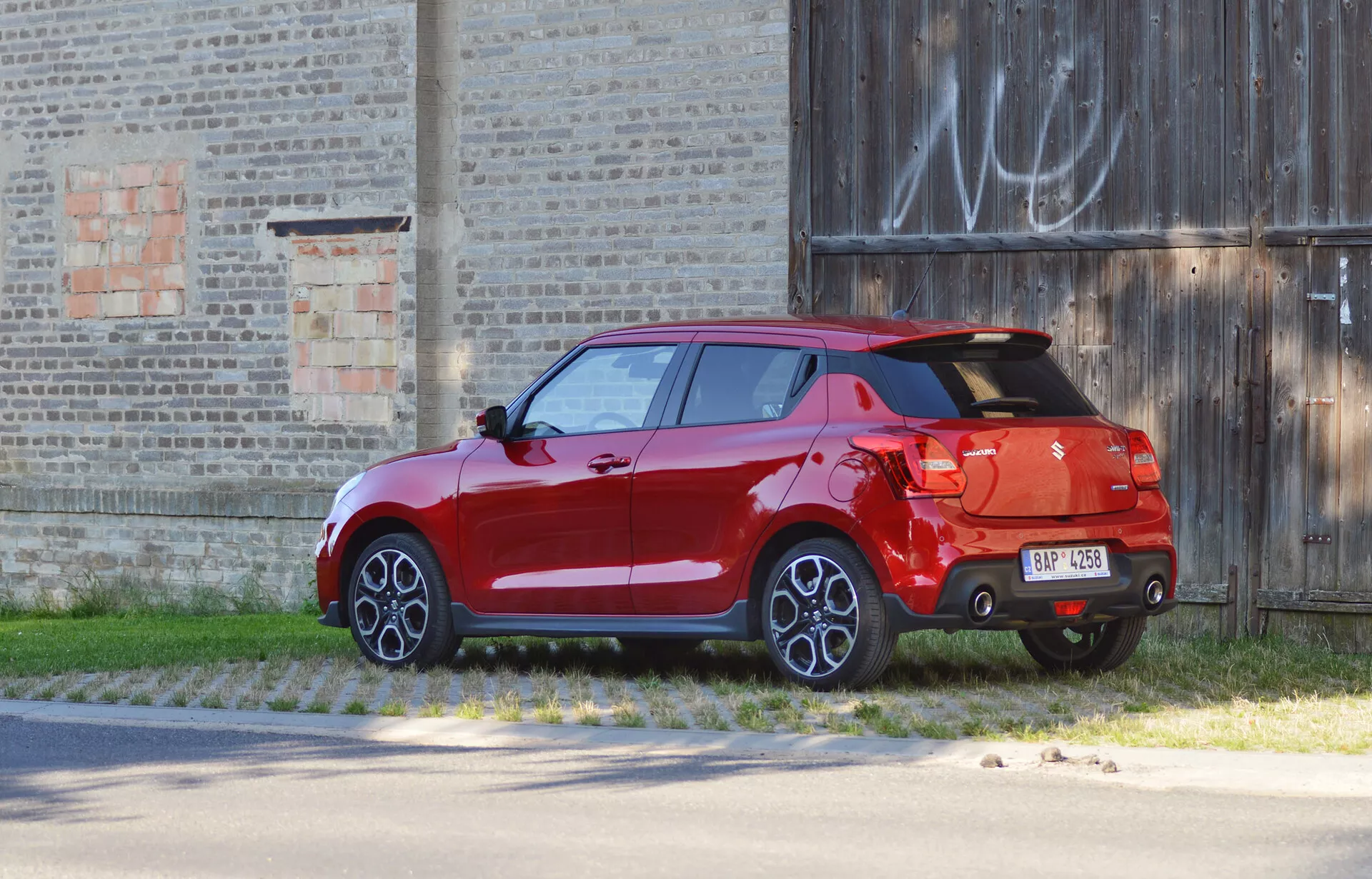 Suzuki Swift Sport 2017 hatchback v jasnej červenej farbe, zachytený z trojštvrťového pohľadu zozadu, zaparkovaný proti starému budovu. Obrázok zdôrazňuje dizajnové prvky, ako sú zadné svetlá, zadný kufor, výrazný zadný spojler, dvojfarebný nárazník s difúzorom, jediná viditeľná výfuková koncovka, viacramenné zliatinové kolesá, úplne namaľovaná karoséria vrátane bočných zrkadiel a dverí, čo naznačuje vyššiu úroveň výbavy alebo luxusné možnosti.