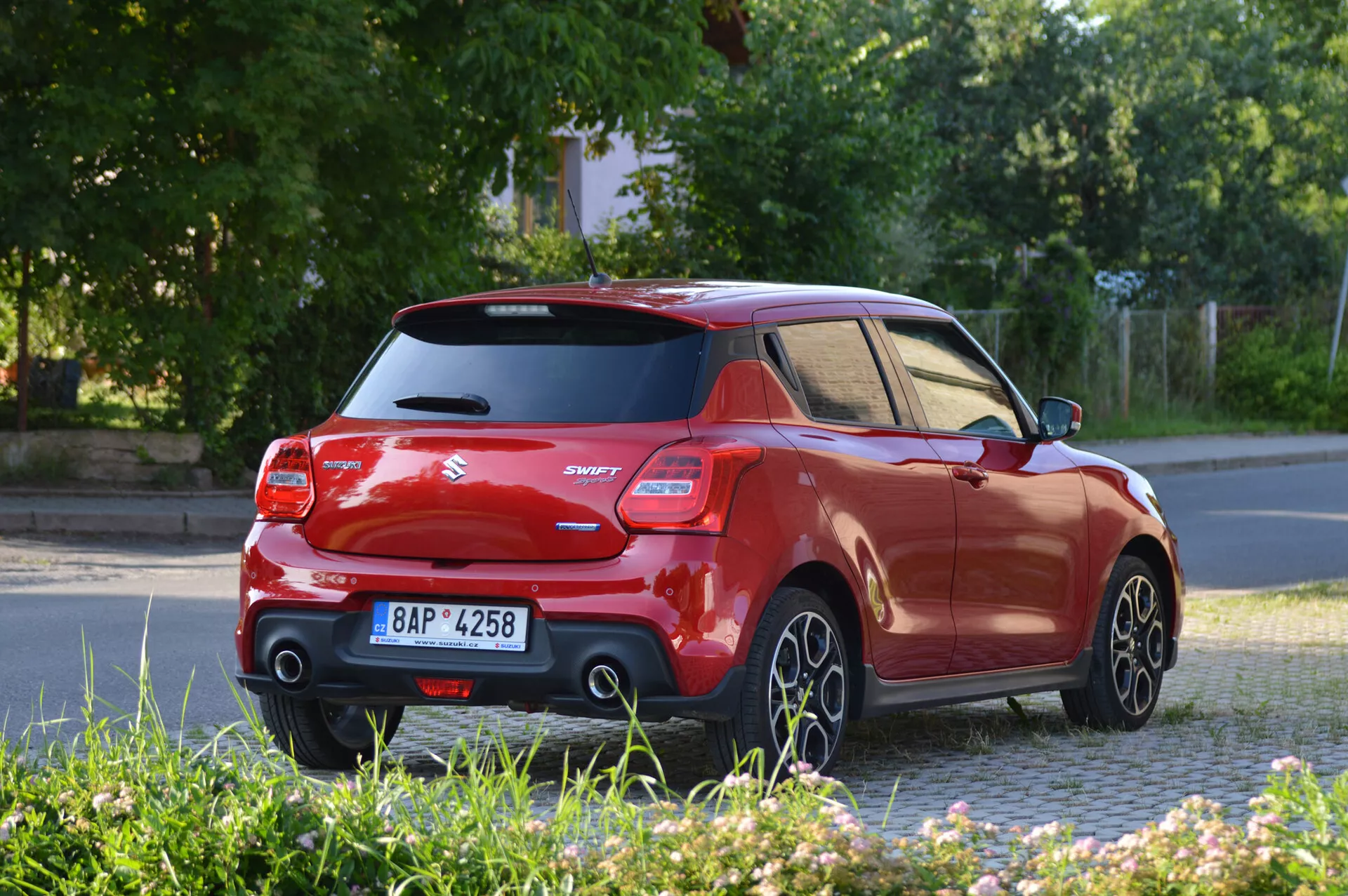 Obrázok predstavuje moderný hatchback Suzuki Swift Sport 2017 zachytený z uhla zadnej tretiny, zobrazujúci bok a zadnú časť vozidla. Výrazné sú kontrastná čierna strecha a stĺpiky, zvýšený zadný spojler, charakteristické zadné svetlá a modelový znak 'Swift'. Fotografia je urobená z úrovne zeme, zobrazujúca pohybujúce sa päťlúčové kolesá z ľahkej zliatiny, čo naznačuje, že vozidlo je v pohybe. Osvetlenie naznačuje, že je skoro ráno alebo neskoro popoludní, s dlhými tieňmi.