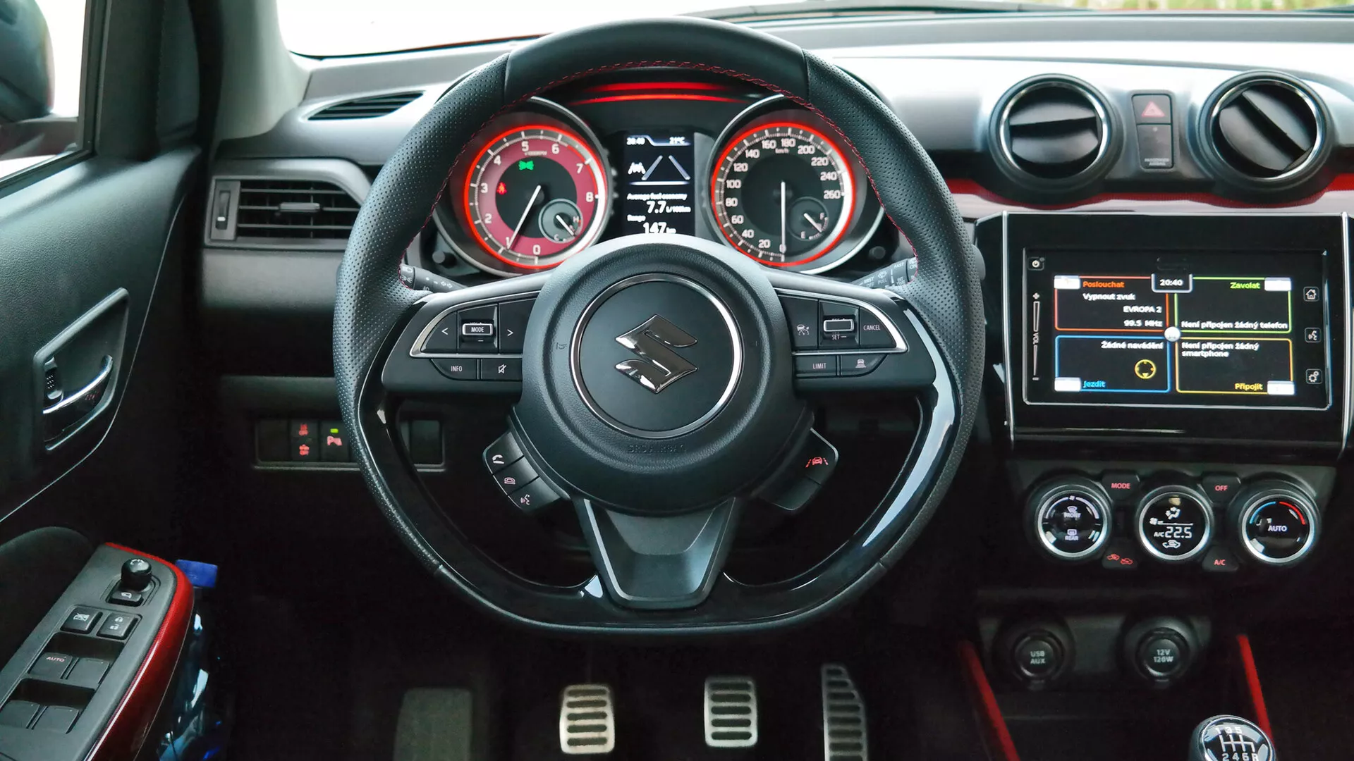 Interiérová fotografia Suzuki Swift Sport 2017 z pohľadu vodiča, s trojramenným volantom s plochým dnom a červeným prešívaním, ovládacími tlačidlami, analógovým otáčkomerom a rýchlomerom s centrálnym digitálnym displejom, dotykovým infotainment systémom, dvojzónovou klimatizáciou s červeným akcentovým osvetlením, kruhovými vetracími otvormi so striebornými akcentmi a držiakmi na nápoje v stredovej konzole.