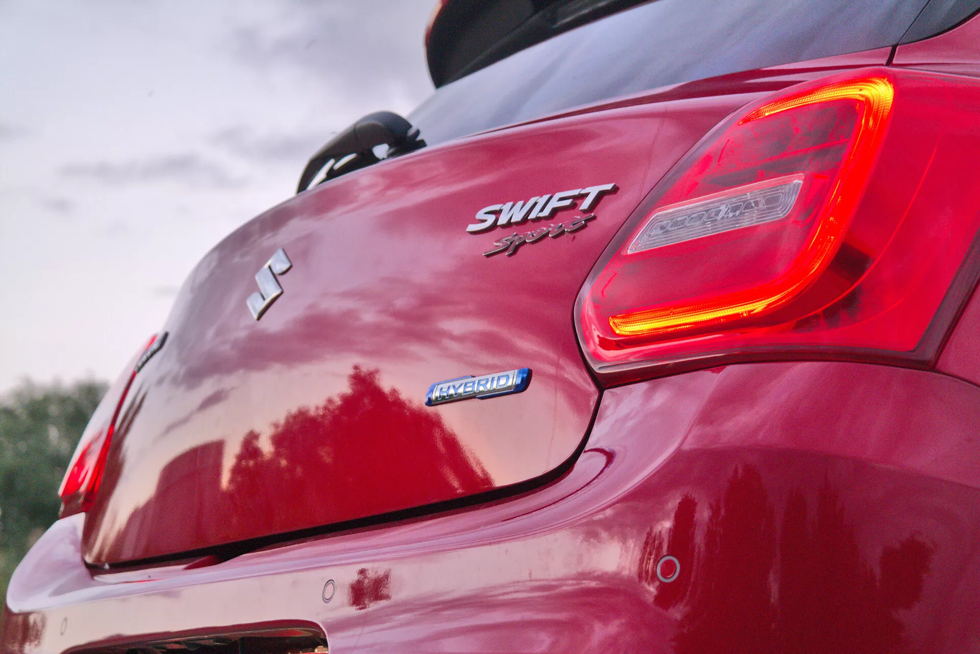 Tento detailný záber zachytáva zadný pravý roh červeného Suzuki Swift Sport 2017, zameraný na označenie auta a osvetlené zadné svetlo. Z nízkeho zadného uhla zvýrazňuje kontúry zadnej časti auta, spoiler a športovú estetiku. Viditeľné prvky zahŕňajú emblém Suzuki 'S', označenie 'SWIFT' a 'Sport', označenie 'HYBRID', dizajn zadného nárazníka, zliatinové kolesá a odrazy naznačujúce lesklý povrch laku.