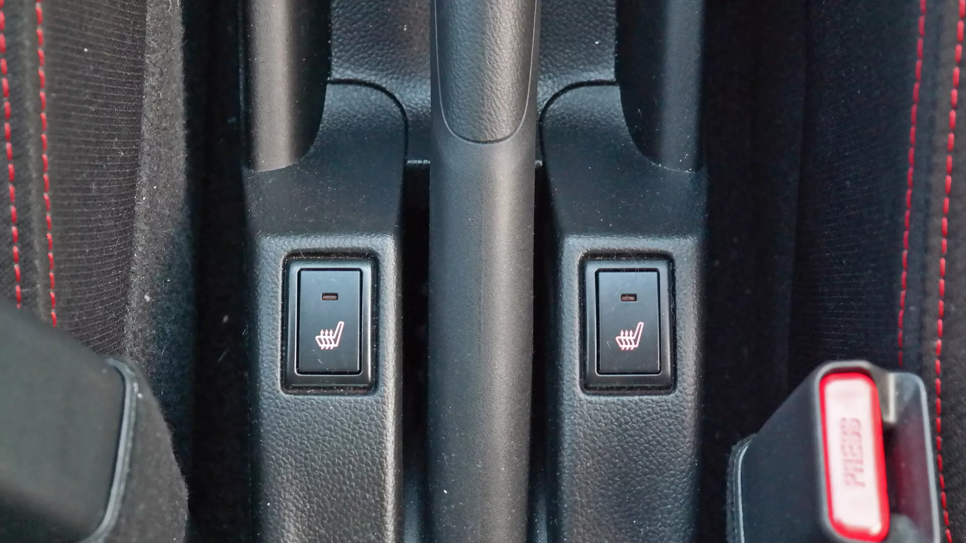 Detailný pohľad na stredovú konzolu interiéru Suzuki Swift Sport 2017 s ovládaním vyhrievaných sedadiel, texturovaným čiernym plastom, červeným prešívaním a čiastočne viditeľnou červenou sponou pásu.