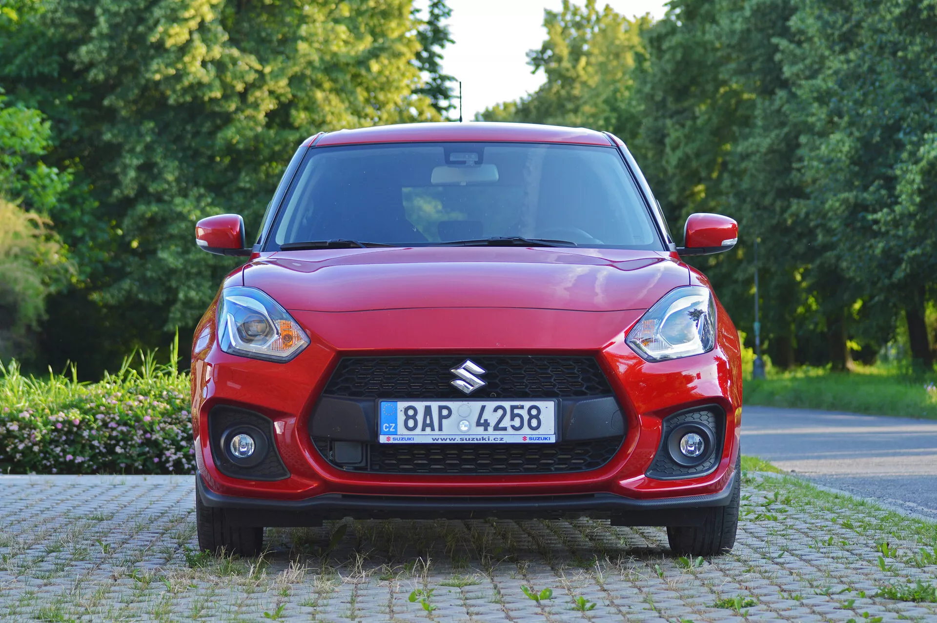 Pohľad z predného uhla na červený hatchback Suzuki Swift Sport 2017 s výraznou mriežkou a modernými svetlometmi, za denného prirodzeného osvetlenia. Kapota vozidla ukazuje aerodynamické línie, zatiaľ čo hmlovky pridávajú na šírke a športovosti. Na čelnom skle je viditeľné možné technologické zariadenie, viacramenné zliatinové kolesá a viditeľná registračná značka. Vozidlo je zaparkované v predmestí alebo parku s vegetáciou.