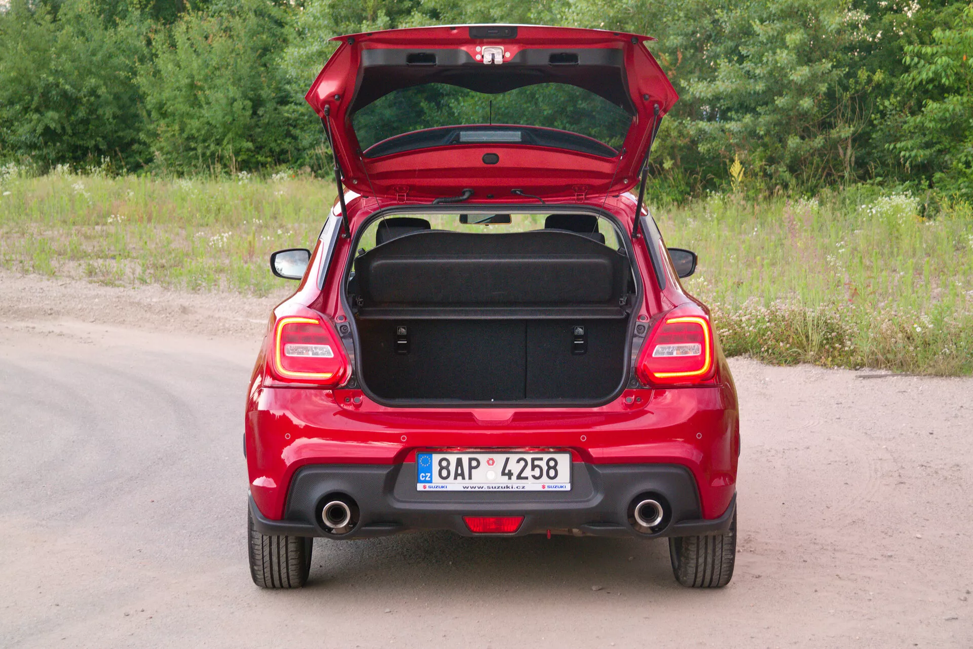 Zadný pohľad na červený hatchback Suzuki Swift Sport 2017 s otvoreným vekom kufra, zobrazujúci symetrické zadné prvky, zadné svetlá, viditeľný výfuk na strane spolujazdca a zadný stierač. Vozidlo je zaparkované na nespevnenom chodníku s trávou a zeleňou v pozadí. ŠPZ je umiestnená centrálne na zadnom nárazníku. Prázdny kufor odhaľuje úložnú kapacitu a vnútorné obloženie s rukoväťou na vnútornej strane veká pre ergonomické použitie. Čiastočne sklopené zadné sedadlá naznačujú 60/40 delenie pre rozšíriteľný nákladový priestor. Jednoduchý zadný nárazník naznačuje štandardné prvky bez pokročilých technologických prídavkov.