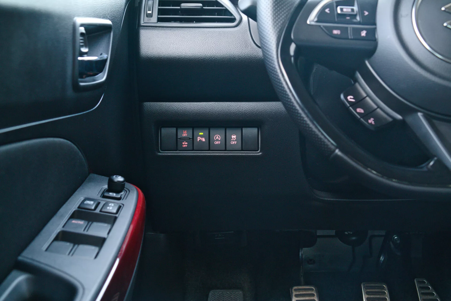 Interiérový pohľad na Suzuki Swift Sport 2017 z pohľadu pasažiera, zobrazujúci prístrojovú dosku vodiča a bočnú časť stredovej konzoly. Zahŕňa čiastočný pohľad na volant s multifunkčnými tlačidlami, prístrojovú dosku s tlačidlami, vodičove dvere s ovládaním elektrických okien a zrkadiel, športové kovové pedále pre manuálnu prevodovku a detailné textúry a materiály.
