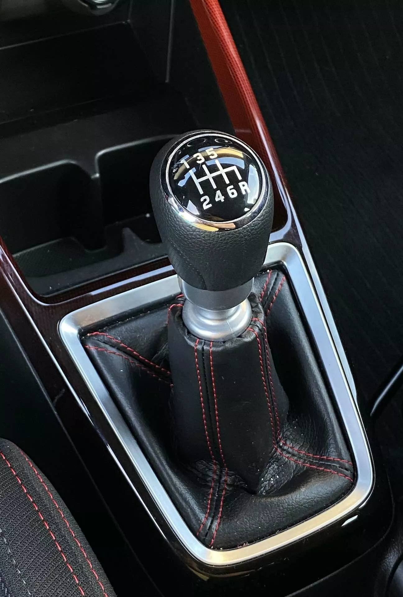 Pohľad zhora a mierne šikmý pohľad na manuálnu radiacu páku v Suzuki Swift Sport 2017, so šesťrýchlostným vzorom ohraničeným bielou farbou na čiernom pozadí. Radiaca páka je umiestnená v stredovej konzole a obklopená koženou manžetou s červeným kontrastným šitím a červenou akcentnou líniou prebiehajúcou paralelne s radiacou pákou. Obrázok ukazuje športový interiér s náznakmi falošného dreveného obloženia, čo naznačuje luxusné dizajnové prvky.