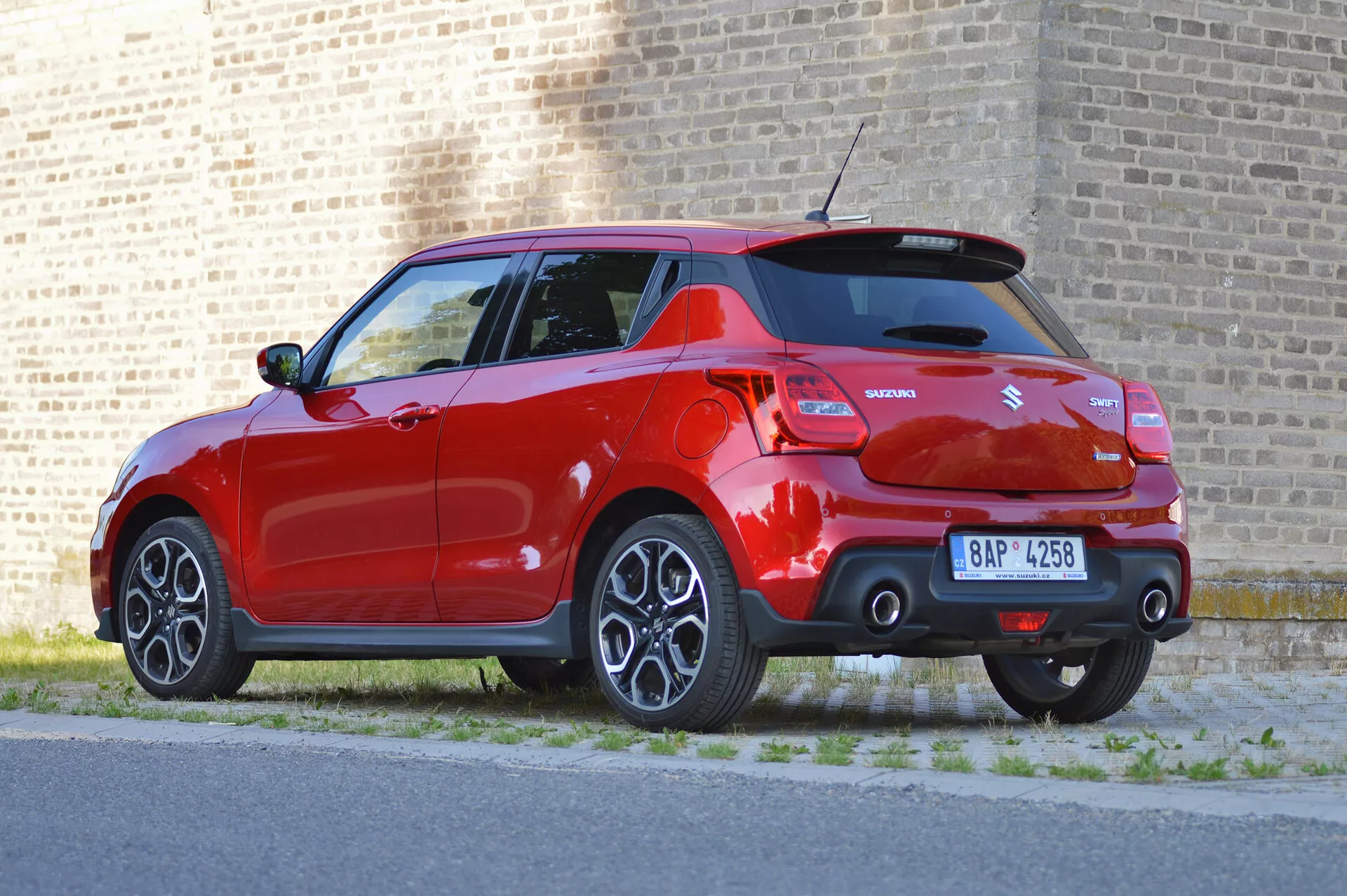Trojštvrtinový zadný pohľad na jasne červený Suzuki Swift Sport 2017, zdôrazňujúci jeho moderný dizajn s prvkami, ako sú zliatinové kolesá, výrazný zadný strešný spojler, dvojité výfuky, krátka rádio anténa, parkovacie senzory a viditeľné značenie. Auto je zaparkované vedľa chodníka s tehlovou stenou v pozadí, za denného svetla.