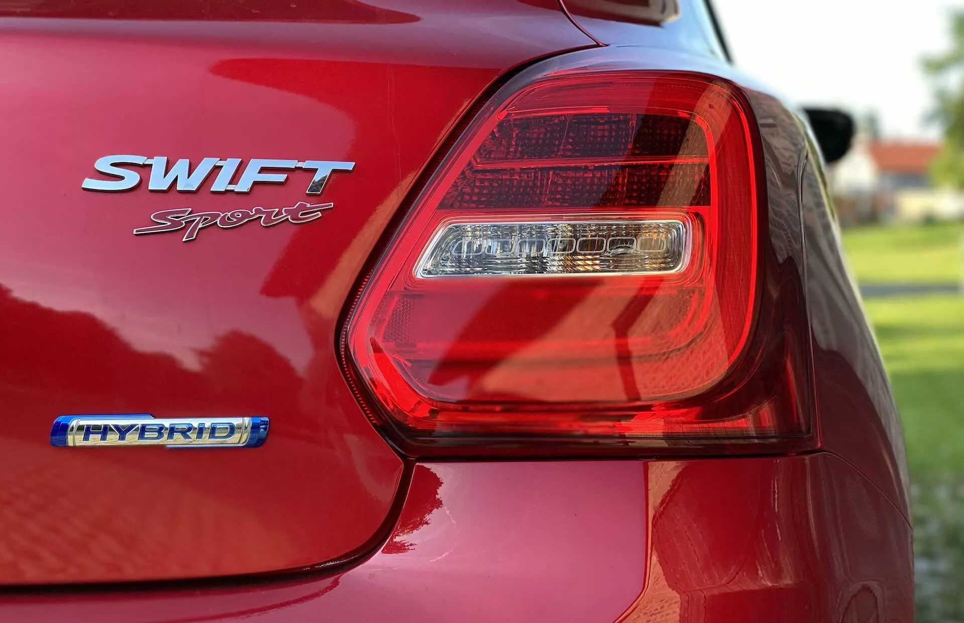 Detailný pohľad na zadnú časť červeného Suzuki Swift Sport 2017, zameraný na ľavé LED zadné svetlo. Obrázok zachytáva lesklý povrch laku, odznaky SWIFT Sport a HYBRID a kontúry zadného blatníka a časti veka kufra. Malá hĺbka ostrosti rozmazáva pozadie a zvýrazňuje detaily svetelného zoskupenia a odznakov, čím zdôrazňuje moderný dizajn a hybridnú technológiu auta.