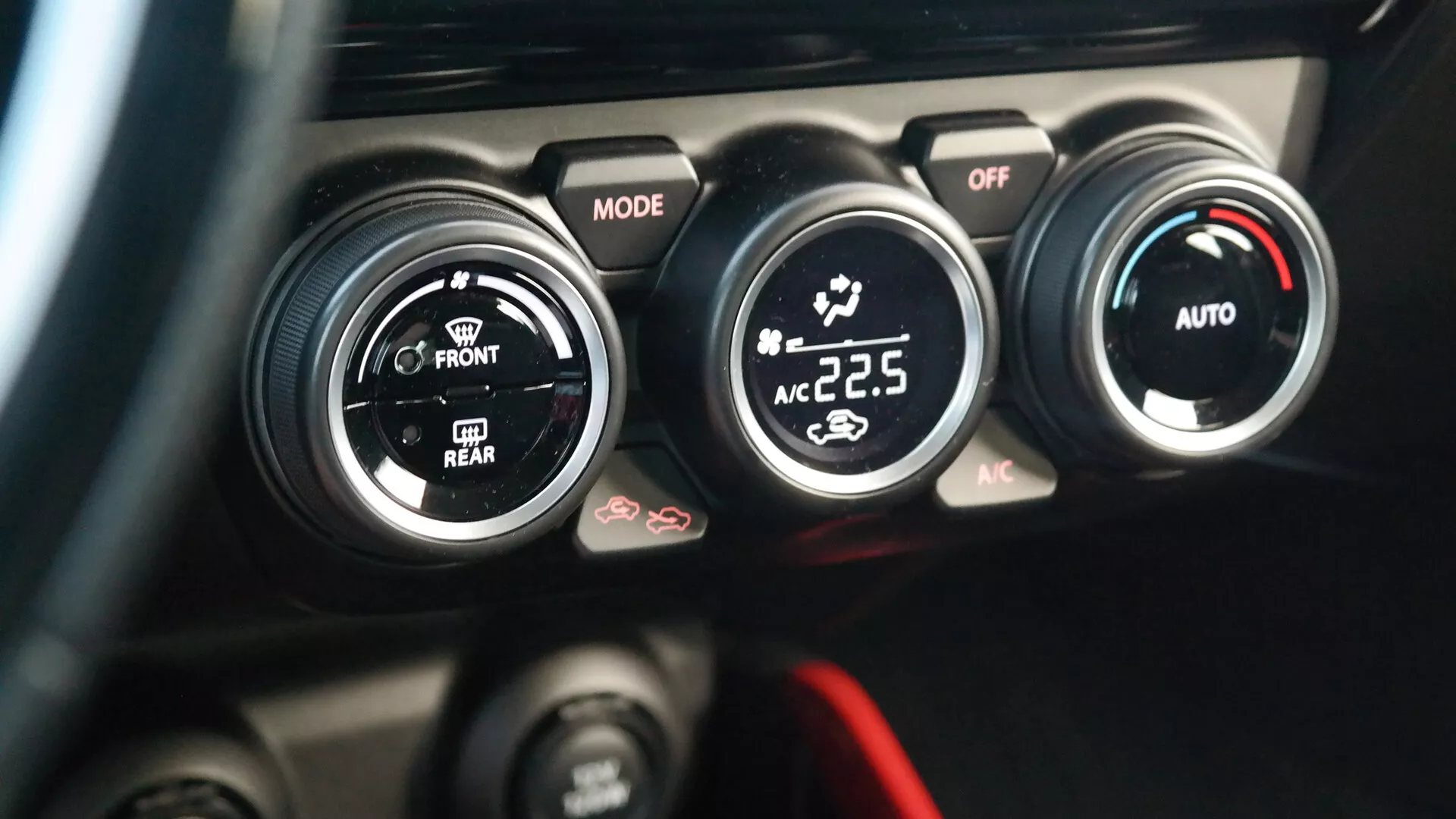 Detailný pohľad na klimatizačný systém Suzuki Swift Sport 2017 z pohľadu vodiča. Obrázok zdôrazňuje stredovú konzolu s tromi čiernymi kruhovými ovládačmi s bielymi a červenými akcentmi; ľavý ovládač označený 'MODE' riadi smer prúdenia vzduchu, stredný ovládač nastavuje výkon a rýchlosť ventilátora s pozíciou 'OFF' a pravý ovládač zobrazuje digitálny údaj o teplote '22,5' stupňa Celzia, čo naznačuje automatickú klimatizáciu s funkciami 'AUTO' a 'AC'. Pod týmito ovládačmi sú čiastočne viditeľné ďalšie tlačidlá, vrátane jedného so symbolom odhŕňania. Pozadie je rozmazané, aby zvýraznilo ovládanie klimatizácie.