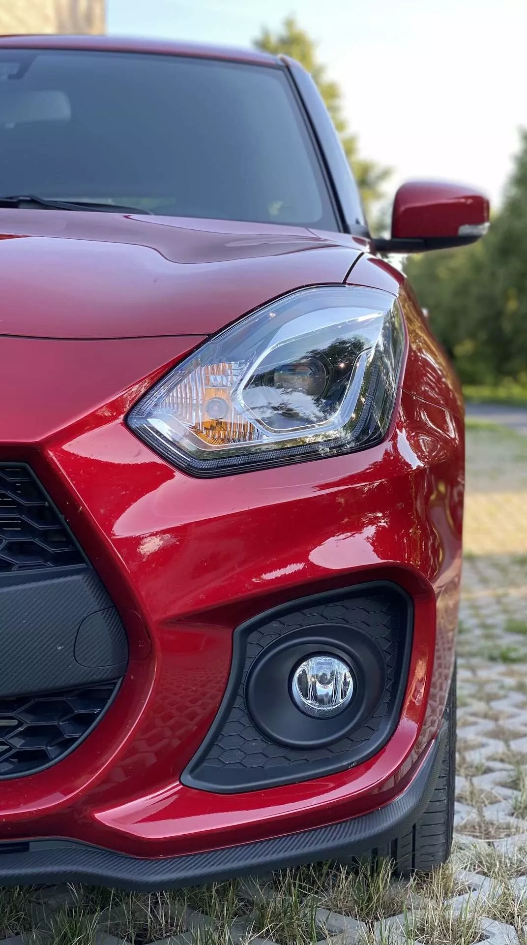 Záber zblízka na červený Suzuki Swift Sport 2017 z nízkeho uhla, zameraný na prednú ľavú časť, zobrazujúci vodičovo svetlomet s prepracovaným dizajnom, potenciálne zahŕňajúcim LED alebo halogénovú technológiu, čiastočne rozmazané logo na mriežke a aerodynamické spätné zrkadlo na pozadí s prírodnou zeleňou.