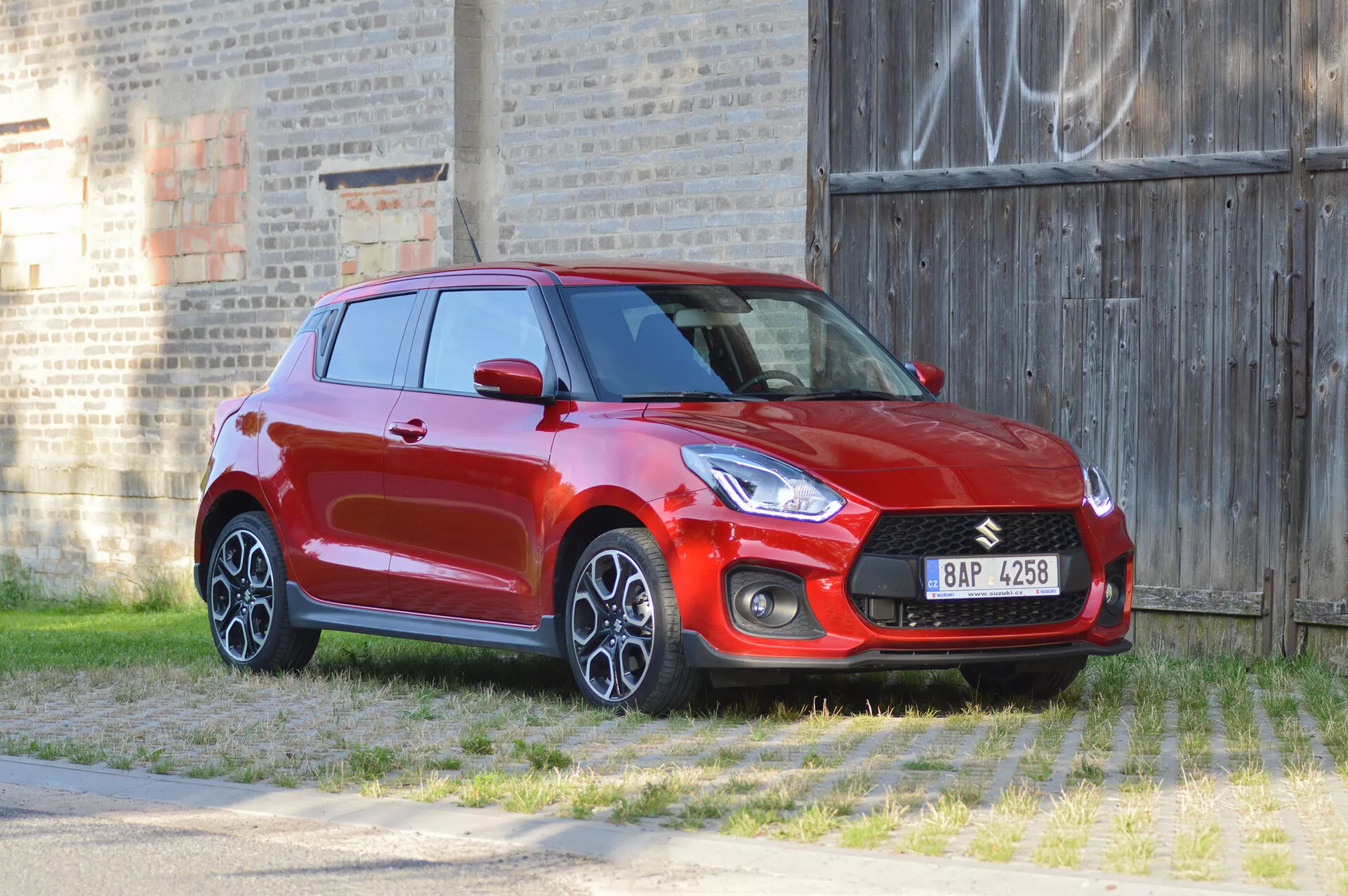 Obrázok červeného hatchbacku Suzuki Swift Sport 2017 zachyteného zo trojštvrťového predného uhla, zvýrazňujúceho predný a bočný profil. Dizajnové prvky vozidla zahŕňajú výraznú prednú masku s integrovaným logom, elegantné svetlomety, kryty hmlových svetiel a viacramenné zliatinové kolesá. Záber je urobený z miernej výšky a poskytuje pohľad zhora na kapotu a strechu. Okolie zahŕňa trávnaté popredie a tehlovú stenu s graffiti v pozadí, čo zdôrazňuje športový a dynamický vzhľad vozidla.