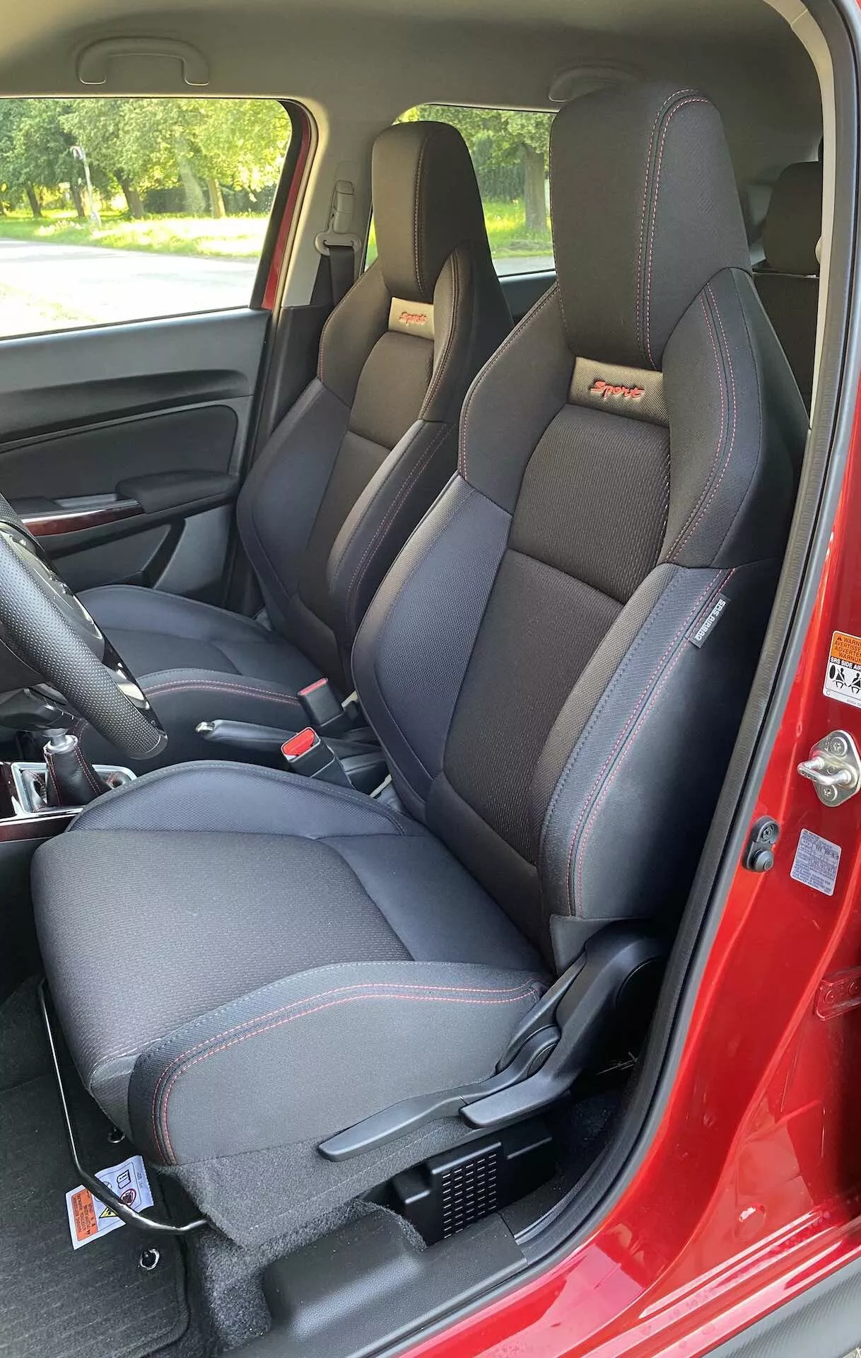 Interiér Suzuki Swift Sport 2017 je zobrazený z perspektívy spolujazdca otvorenými prednými dverami. Zameranie je na moderné, športové tvarované predné sedadlá so vstavanými opierkami hlavy a výraznými bočnými oporami, pravdepodobne s kombináciou koženky a textilu a kontrastným prešívaním. Na hornej časti operadiel sú viditeľné embosované alebo vyšívané logá. Obidve sedadlá sú vybavené trojbodovými bezpečnostnými pásmi, pričom na strane vodiča je nastaviteľný upevňovací mechanizmus pre nastavenie výšky pásu na stĺpiku B. Stredová konzola medzi sedadlami má úložné priestory a páku ručnej brzdy. Tmavý interiérový panel dverí obsahuje kľučku dverí, ovládanie nastavenia zrkadiel a elektrického ovládania okien. Podlaha je pokrytá tmavým kobercom a základňa sedadiel ukazuje montážne koľajnice. Denné svetlo a prírodné vonkajšie prostredie sú viditeľné cez okná, čo poskytuje odrazy a tiene, ktoré zvýrazňujú detaily interiéru.