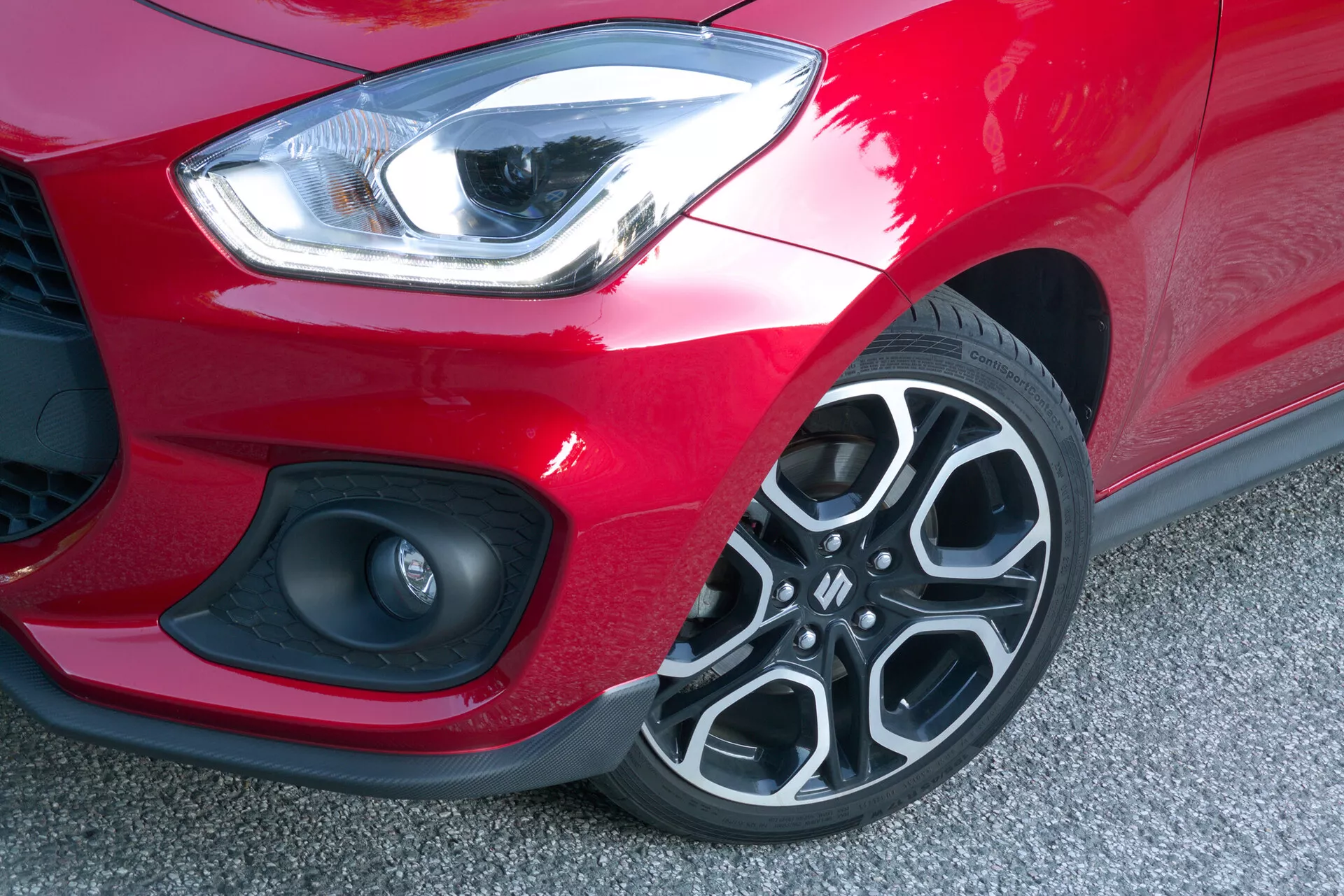 Detailný záber na predný ľavý roh modelu Suzuki Swift Sport z roku 2017, odfotený z uhla zhora a mierne spredu. Elegantný, zakrivený LED svetlomet vyžaruje jasné biele svetlo, čo naznačuje funkciu denného svietenia. Pod svetlometom je viditeľná malá časť mriežky so vzorom sieťky vedľa kruhového prvku, ktorý môže byť hmlovým svetlom alebo parkovacím senzorom, umiestneným v dynamicky tvarovanom prednom nárazníku. Matná čierna časť nárazníku kontrastuje so žiarivo červeným lakom auta. Výrazný viaclúčový dizajn zliatinových kolies s tmavými a striebornými prvkami a viditeľným vzorom pneumatiky naznačuje dobrý stav. Nízká výška auta od zeme je zreteľná, čo odzrkadľuje štandardnú výšku vhodnú pre športový model.