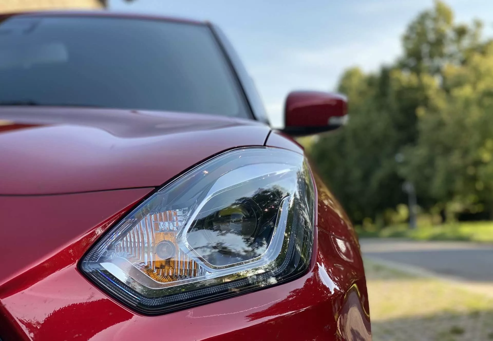 Detailný záber predného ľavého svetlometu vozidla Suzuki Swift Sport 2017. Obrázok je vytvorený z predného šikmého pohľadu, ktorý zdôrazňuje detaily dizajnu číreho svetlometu s jeho zložitým vnútorným reflektorom a oranžovým ukazovateľom smeru. Viditeľné sú lesklé, tmavočervené karosérie vrátane kapoty a blatníka, s rozmazaným vonkajším pozadím, stromami a jasnou oblohou.