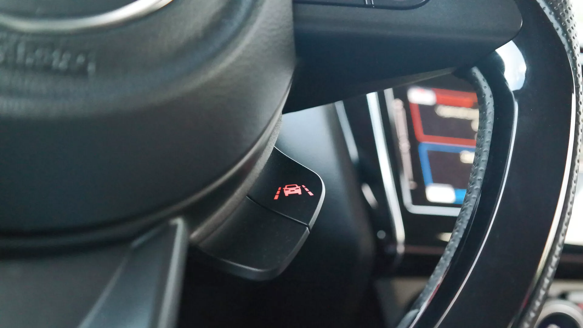 Pohľad na interiér Suzuki Swift 2017 (Swift-sport) z perspektívy sedadla vodiča so zameraním na volant a ovládaciu páku. Tmavý volant, pravdepodobne kožený, má kovový alebo lesklý lem. Ovládacia páka pre tempomat s obrázkom meradla a šípky vyčnieva zo stĺpika riadenia. V rozmazanom pozadí je viditeľný digitálny prístrojový panel zobrazujúci rôzne číslice a čiary, čo naznačuje pokročilé technologické funkcie. Materiály v interiéri kombinujú syntetické alebo kožené textúry so zložitým šitím a plytká hĺbka poľa zvýrazňuje ovládaciu páku s prístrojovou doskou v jemnom rozostrení.